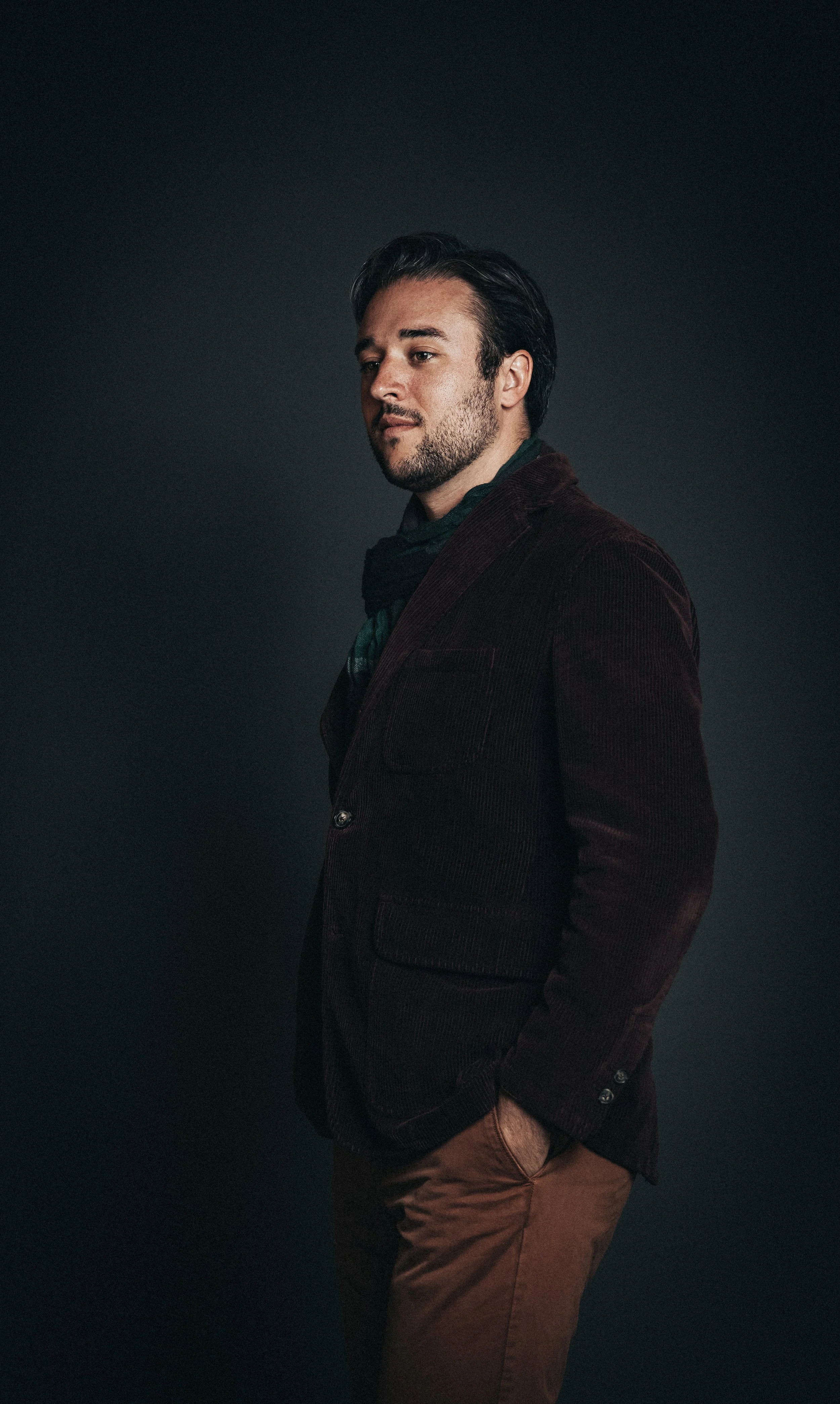A man wearing a dark green shirt, maroon velvet blazer, and brown pants, standing against a dark background with his hands in his pockets, looking slightly to the side.