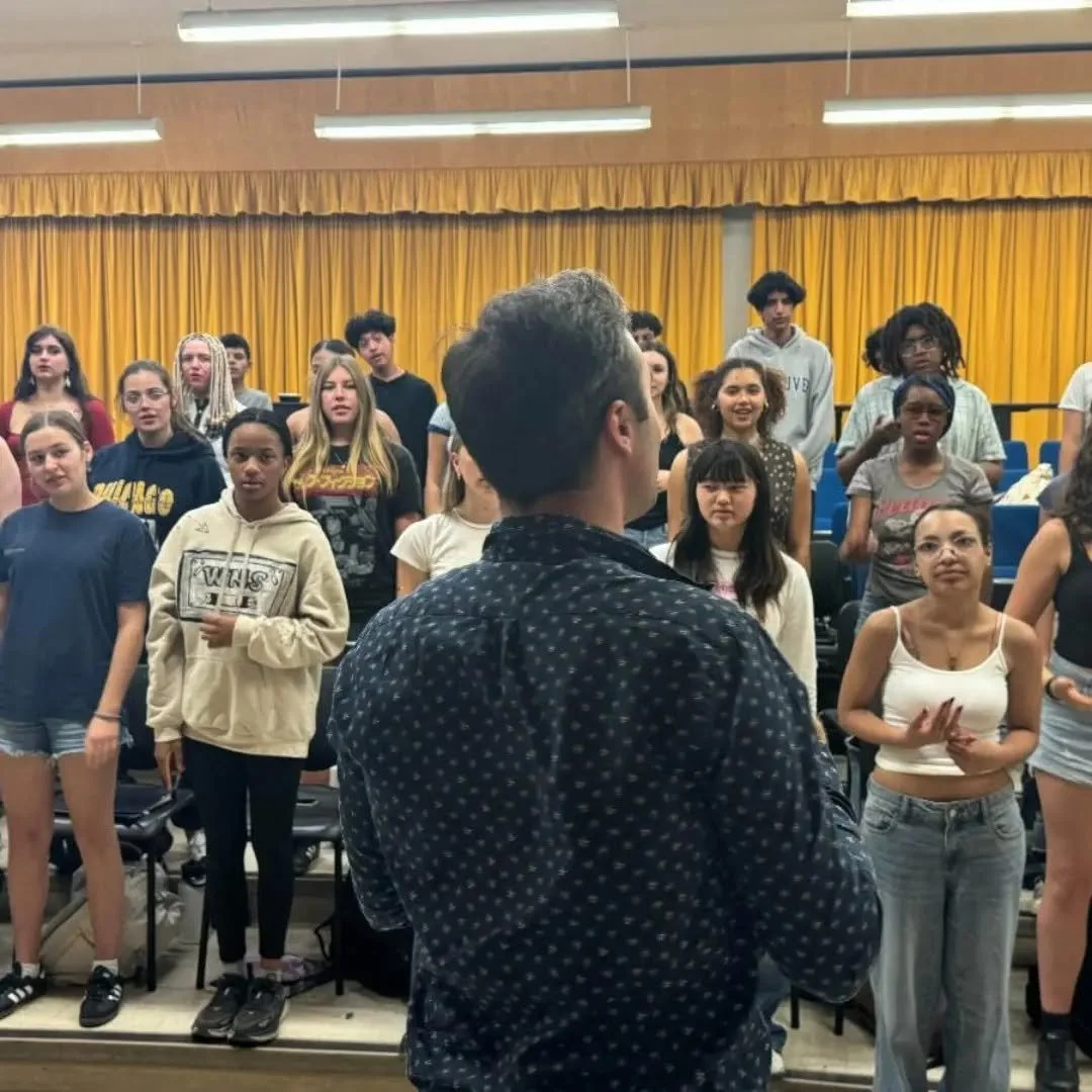 A group of diverse young people standing in an auditorium with yellow curtains, listening to a speaker in the foreground.