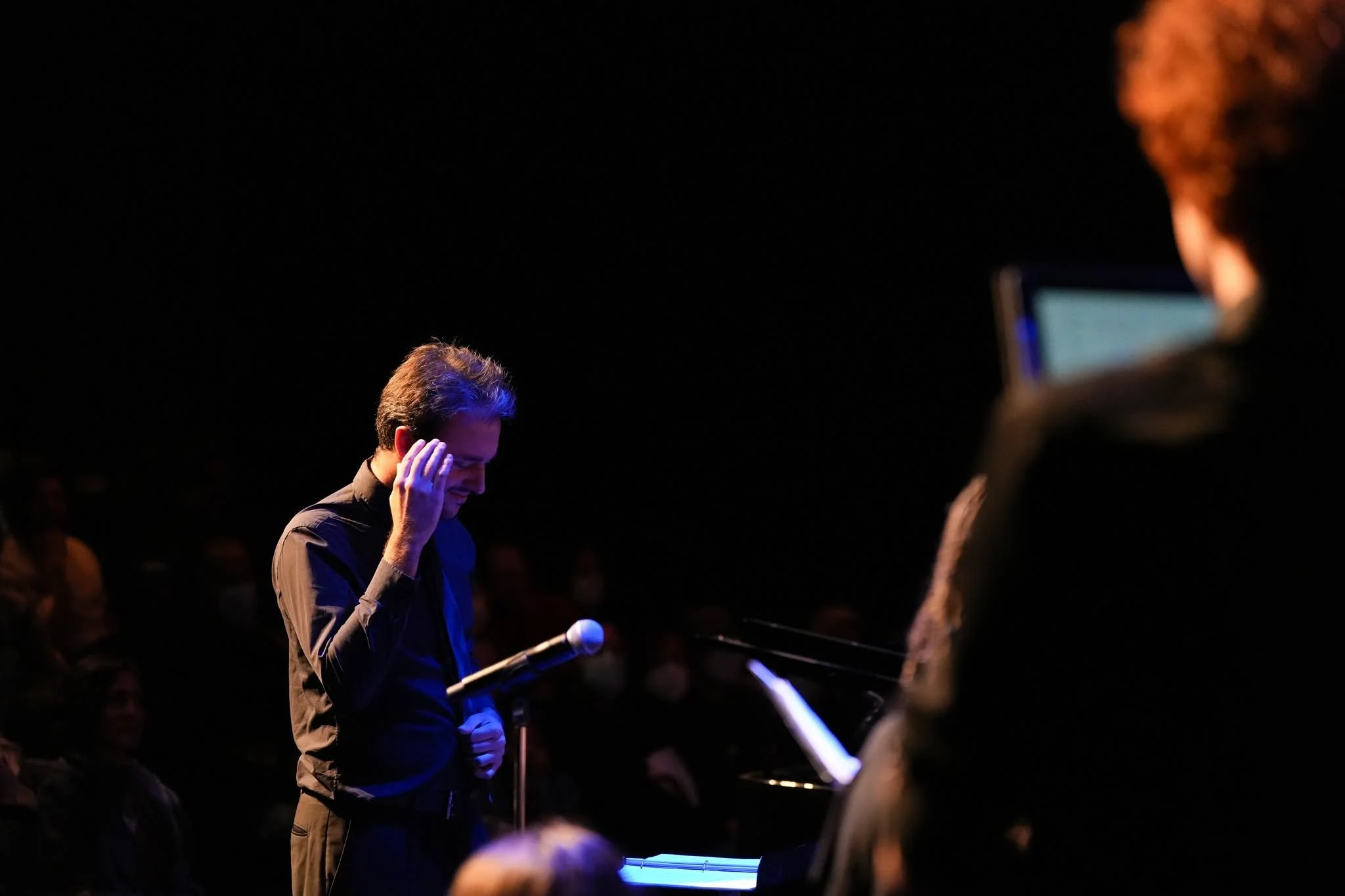 A man in a dark shirt is kneeling with his hand on his forehead, holding a microphone in the other hand, during a performance or event on a dark stage. Another person is partially visible on the right, and a crowd can be seen in the background.