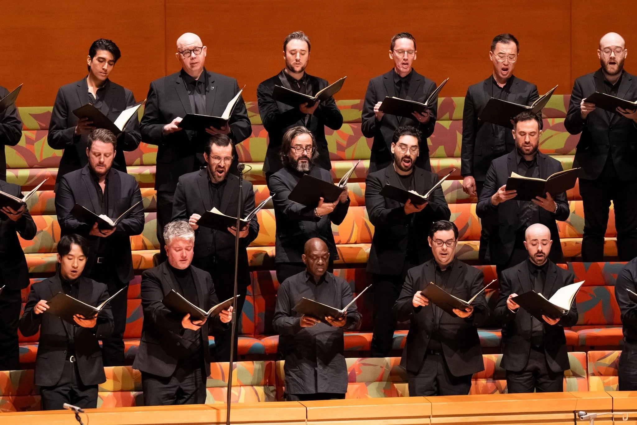A choir of men and women singing during a performance, standing on tiered colorful seating, holding sheet music.