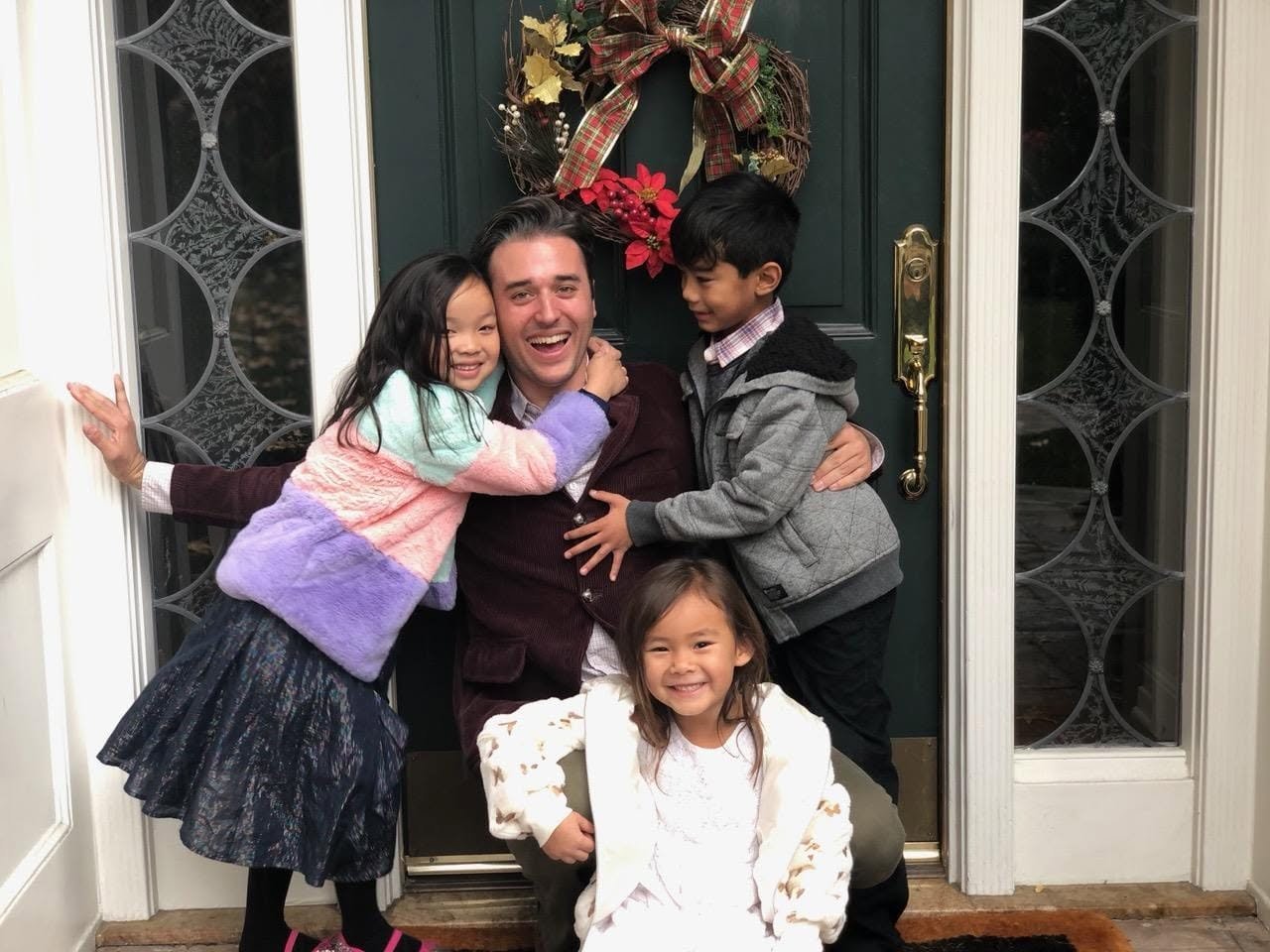 A man and three children hugging and smiling in front of a decorated front door with a holiday wreath.