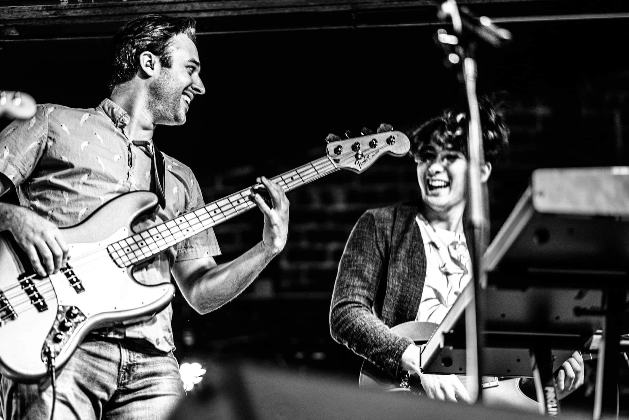 Two musicians performing with one playing a bass guitar and the other playing a keyboard, both smiling.