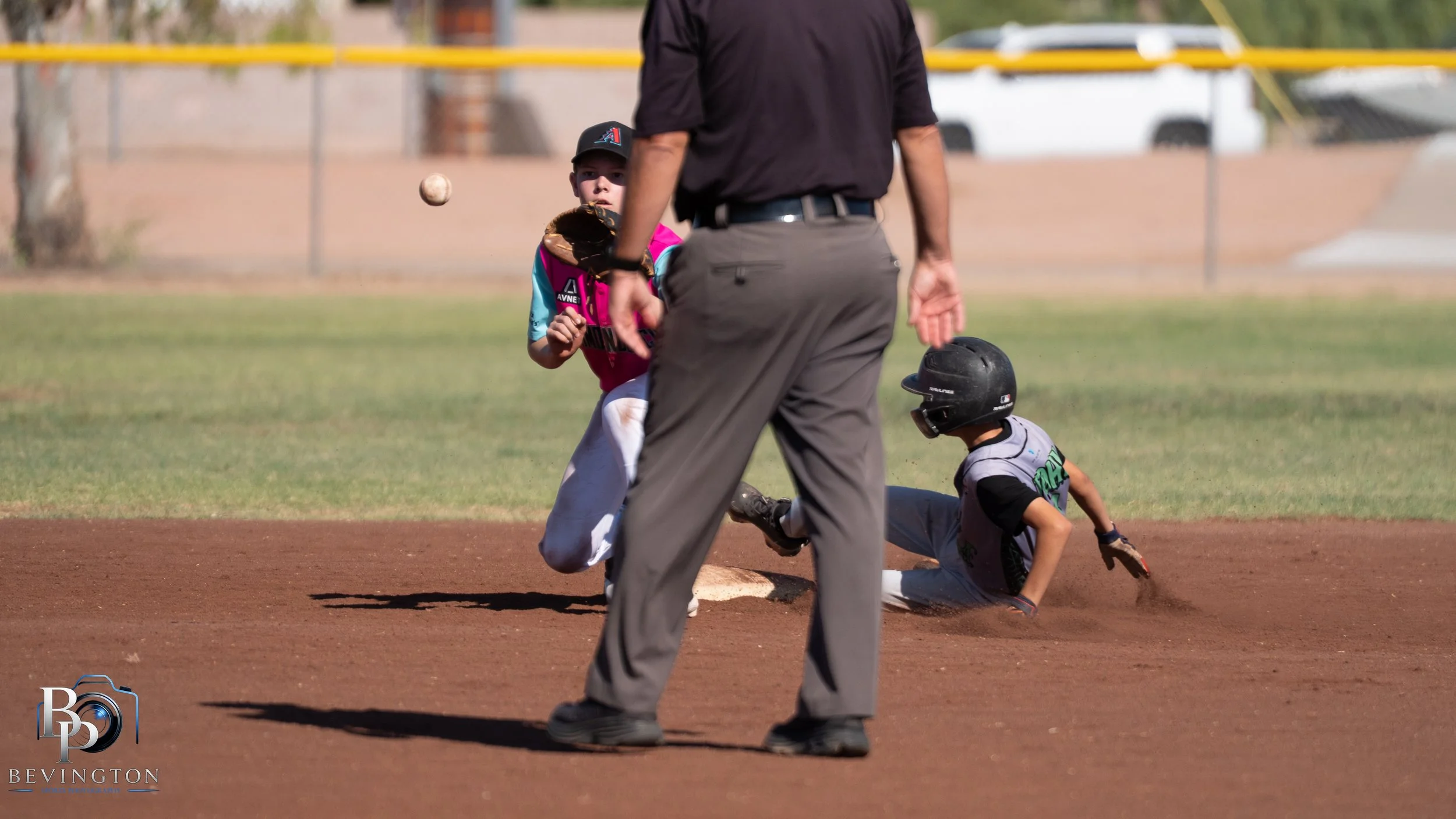 A young baseball player is running toward first base, starting to slide as an opponent attempts to tag him out. An umpire is standing nearby, and another player is on the ground near the base. The scene takes place on a baseball field during daytime.