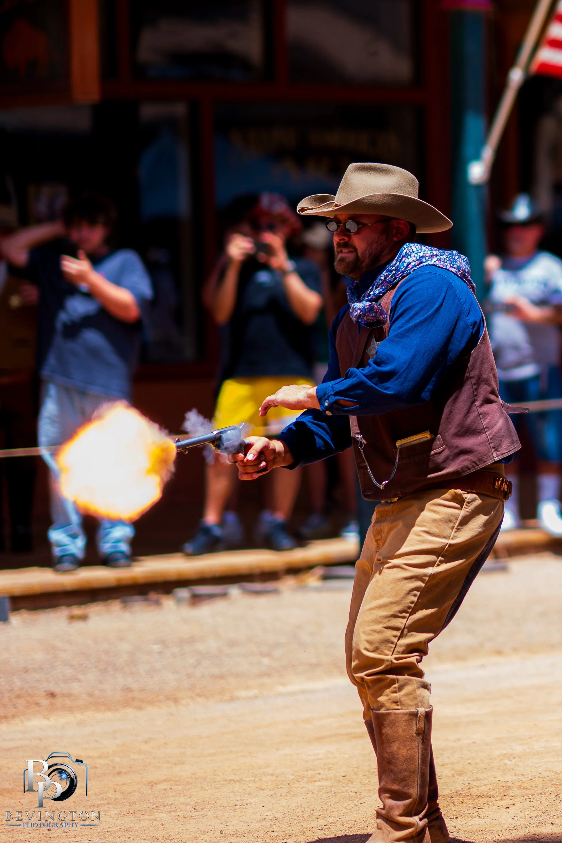 A man in cowboy attire, wearing a wide-brimmed hat, sunglasses, a blue shirt, a patterned bandana, and tan chaps, is firing a gun that produces a visible muzzle flash at a cowboy-themed event. In the background, people are watching and taking photos, with a wood structure and American flag visible.