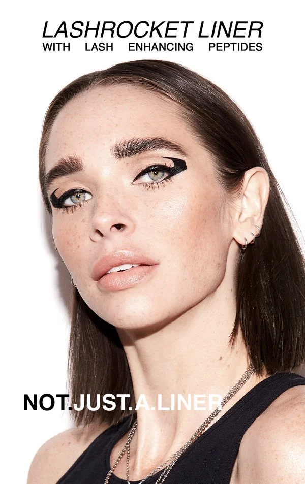 Close-up of a woman with dark brown hair, light skin with freckles, and makeup emphasizing her eyes and lips, promoting Lashrocket Liner with lash enhancing peptides.