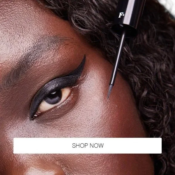 Close-up of a woman’s eye with dramatic black eyeliner and eyeshadow, holding a makeup brush near her face, with a "Shop Now" button overlay.