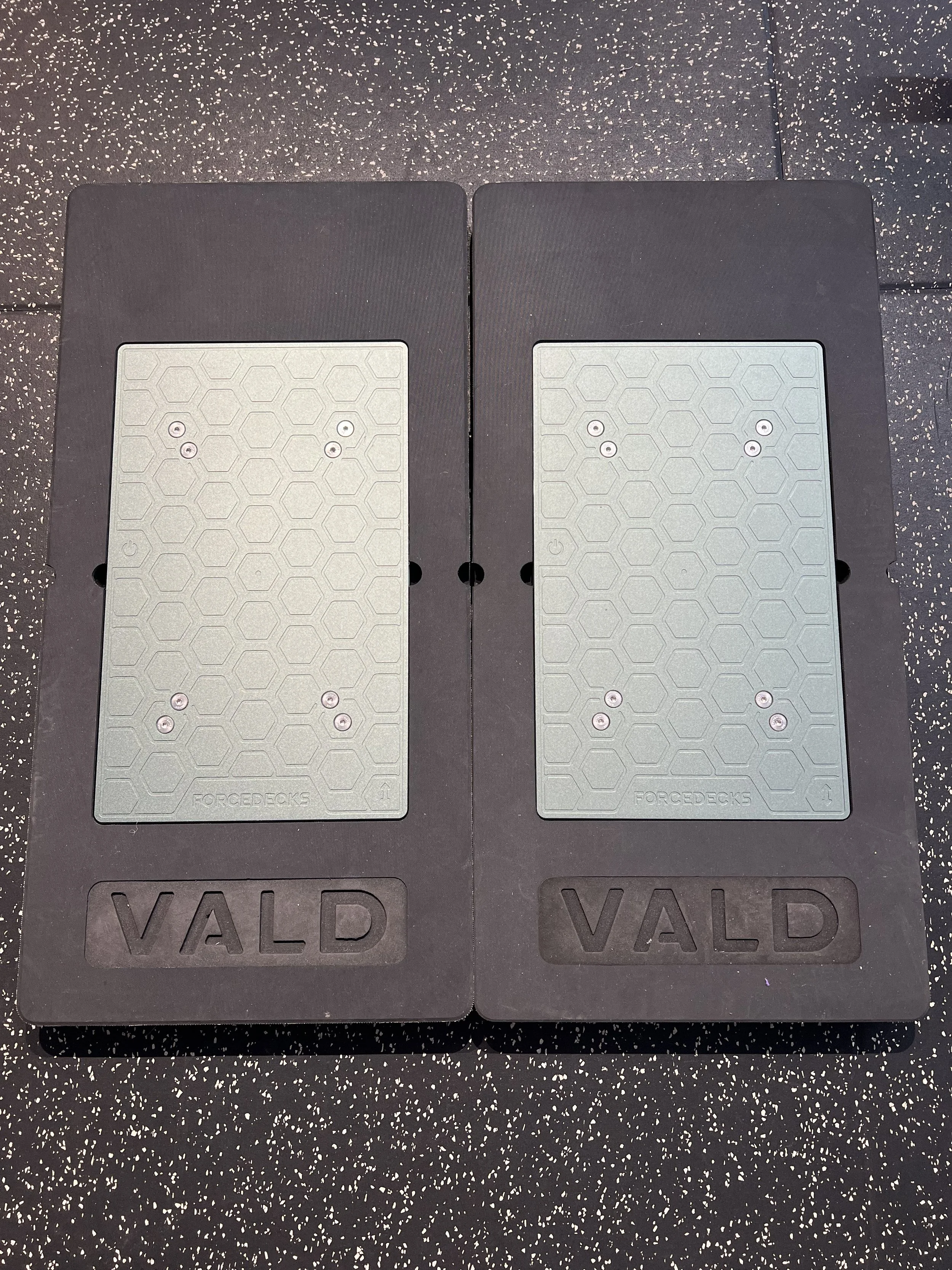 Two black rubber gym step platforms with grey textured surfaces, labeled 'FORCEDECKS' and 'WALD' on black rubber mats on a speckled black and white gym floor.