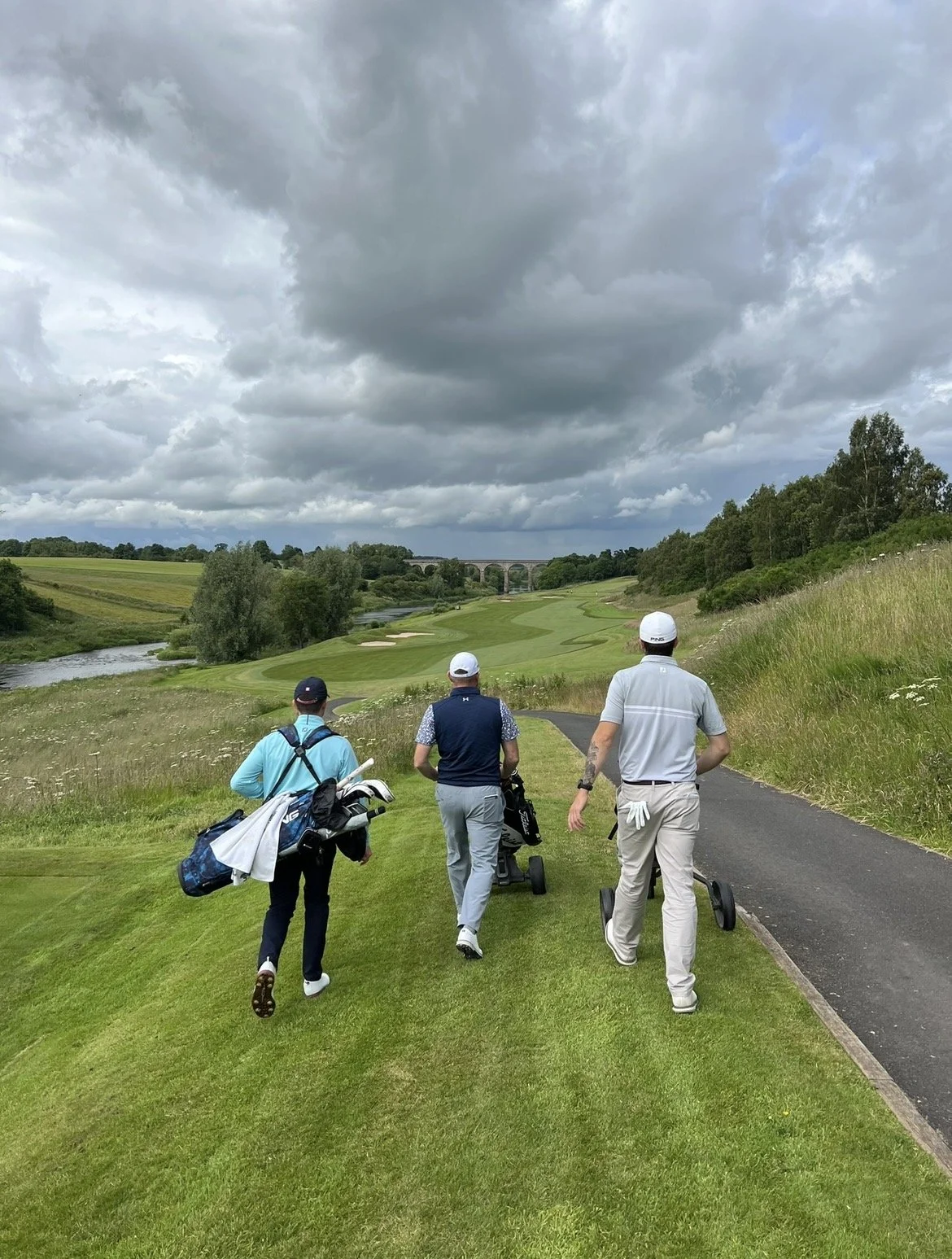 Three men walking on a golf course path, carrying golf bags and equipment, with a scenic view of a river, trees, a bridge, and cloudy sky in the background.