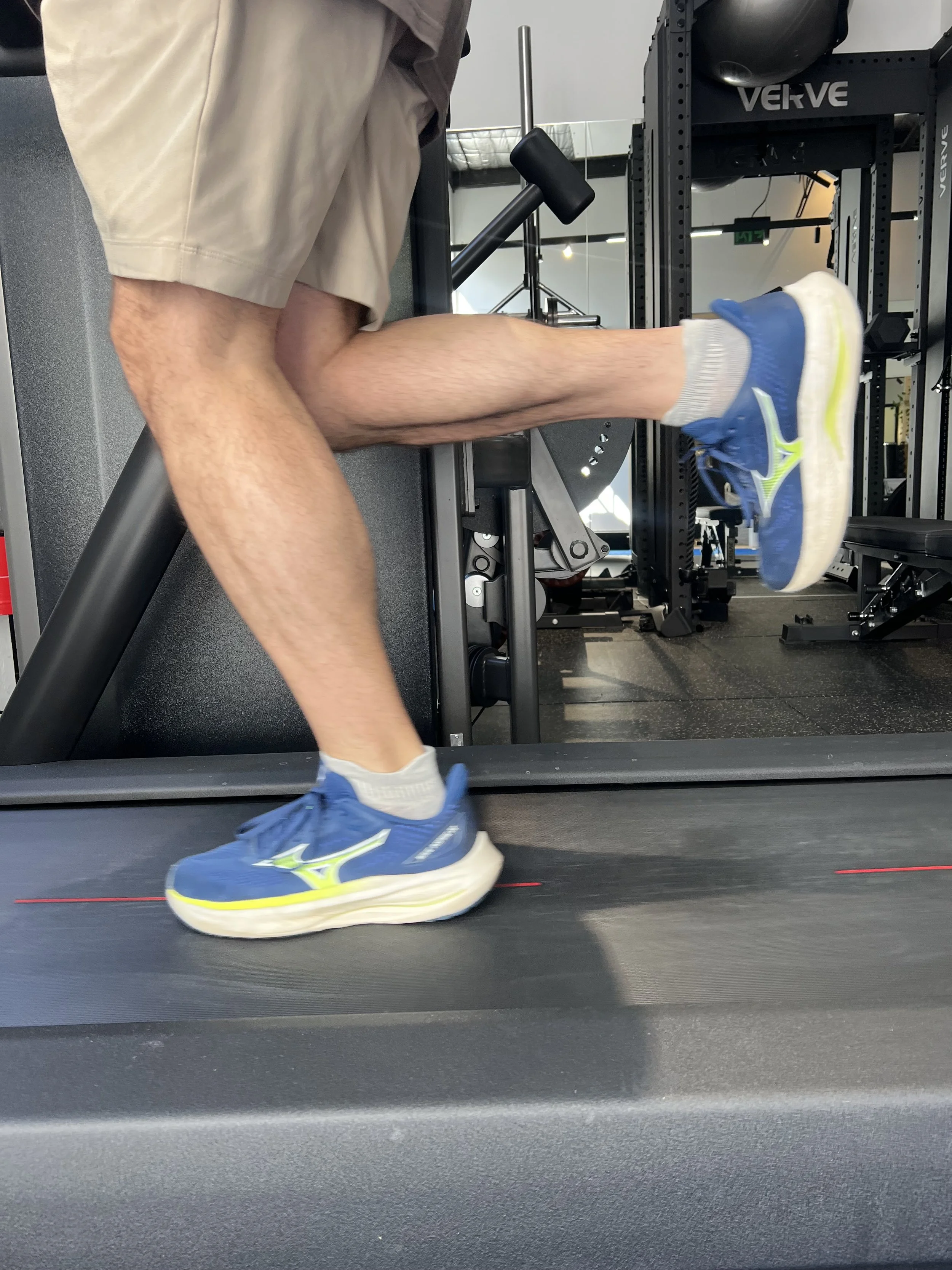 Person running on a treadmill in a gym, wearing blue and white running shoes, shorts, and white socks.