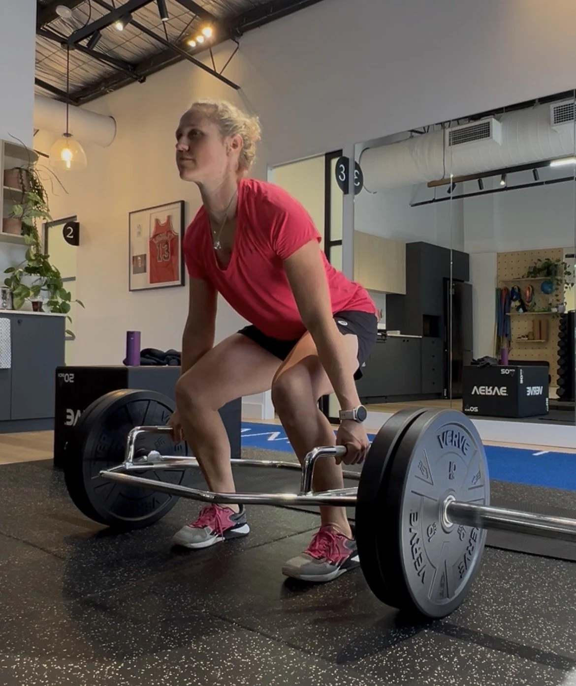 A woman performing deadlifts in a gym, wearing a red shirt, black shorts, and pink athletic shoes.