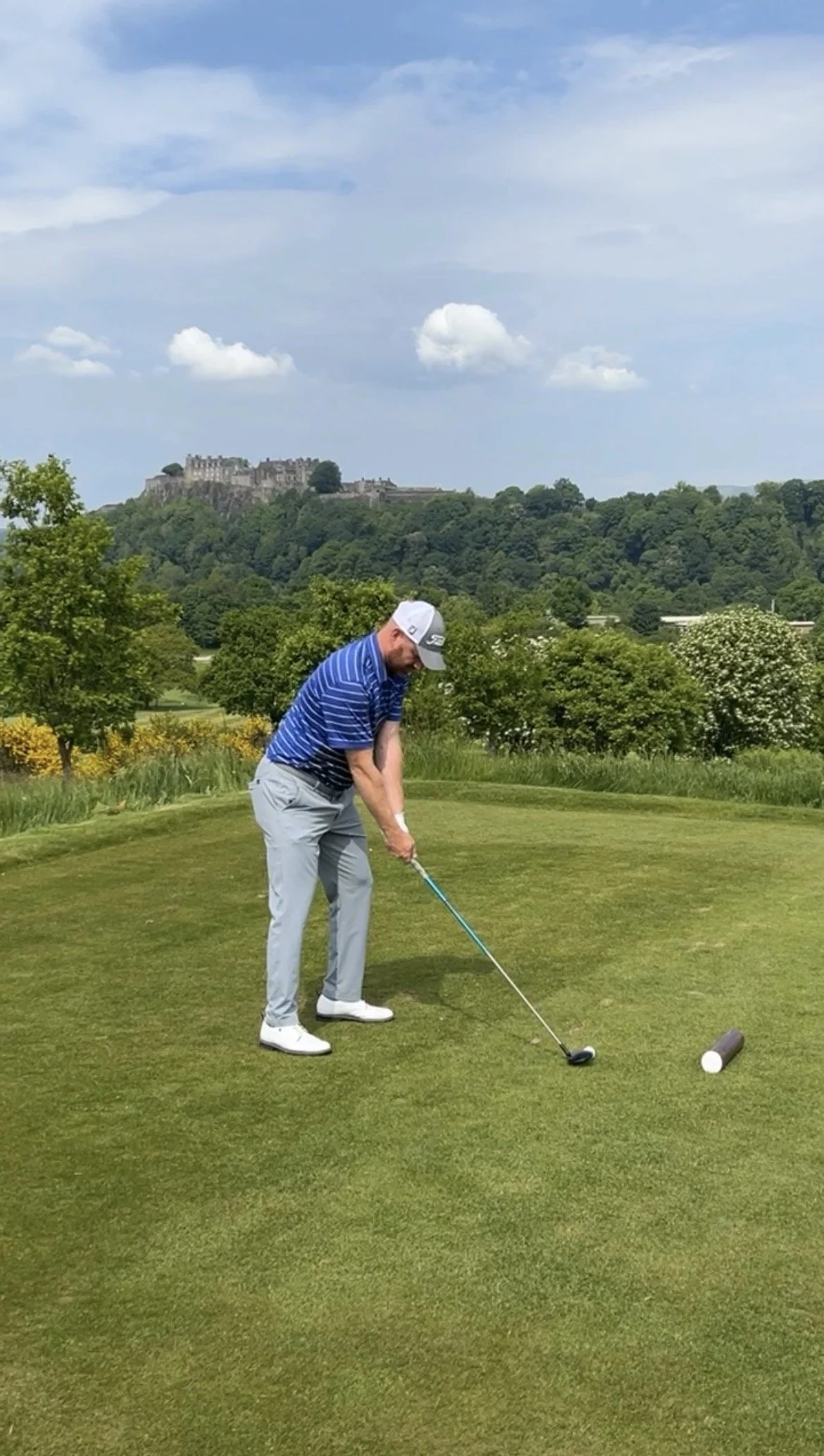 A man in a blue striped polo shirt, light gray pants, white shoes, and a white golf cap preparing to hit a golf ball on a green golf course with trees and a castle on a hill in the background.