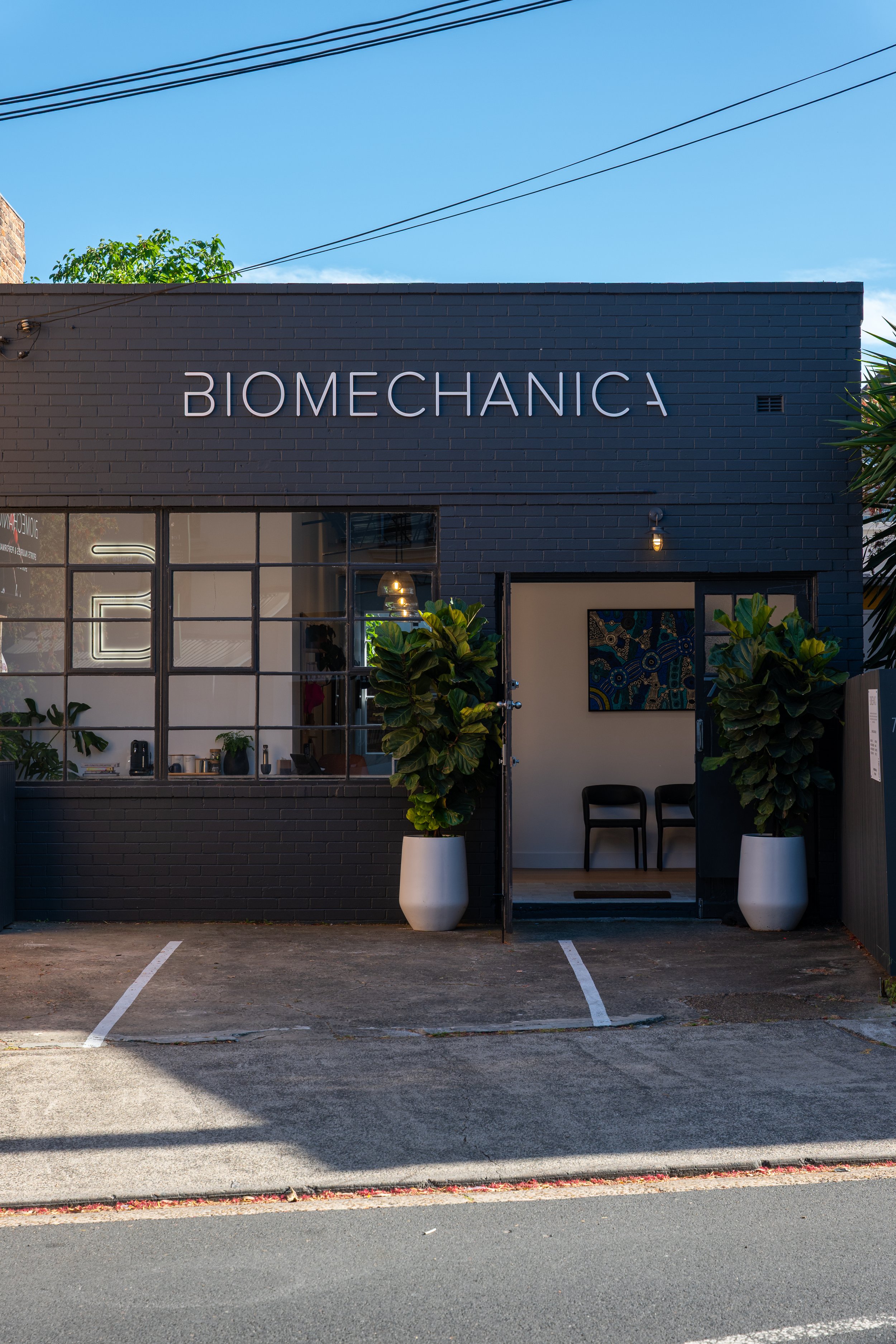 Front of a modern building with a black brick facade and a sign that reads 'BIOMECHANICS' in large white letters. There are large glass windows and a small open entrance with two black chairs inside. Two large potted plants are placed at the entrance, and the parking lot in front has two empty spaces.