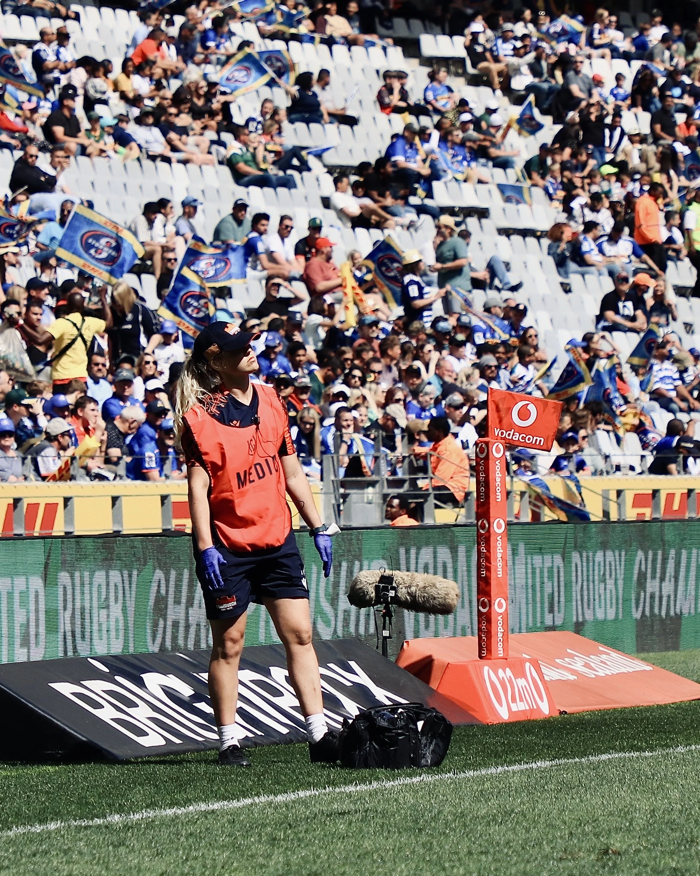 A soccer player in a red jersey and black shorts stands near the sideline during a match, holding gloves. Spectators sit in the background in a stadium, some holding flags and wearing team colors.