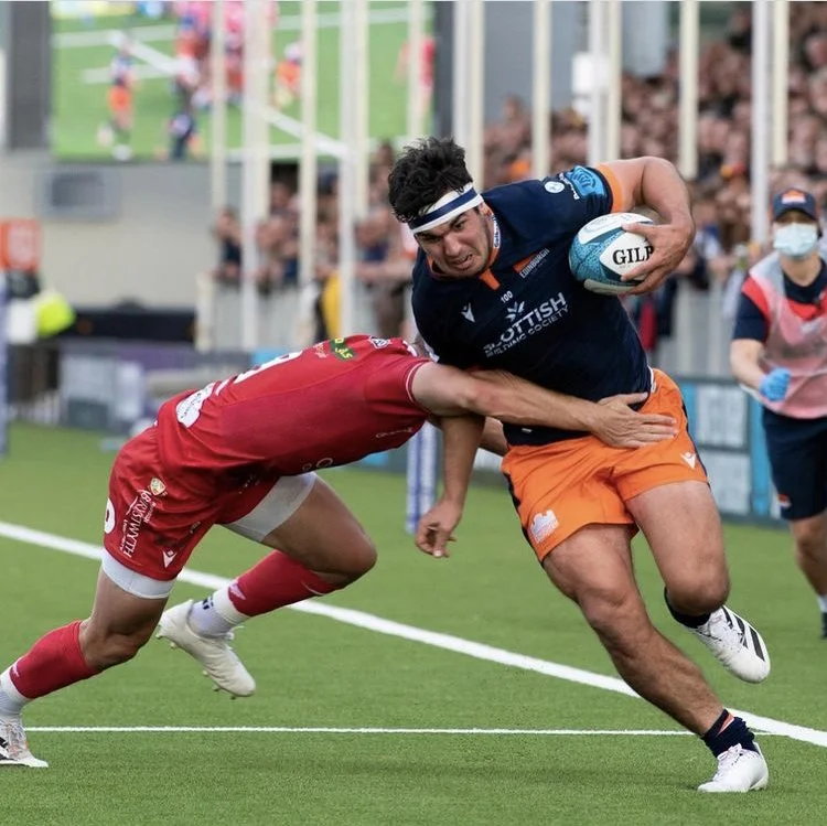 Rugby player in navy blue and orange uniform running with ball while being tackled by player in red uniform on a rugby field.
