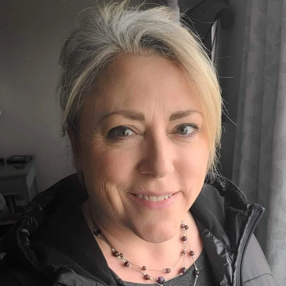 Smiling blonde woman taking a close-up selfie indoors, wearing a black jacket and beaded necklace.