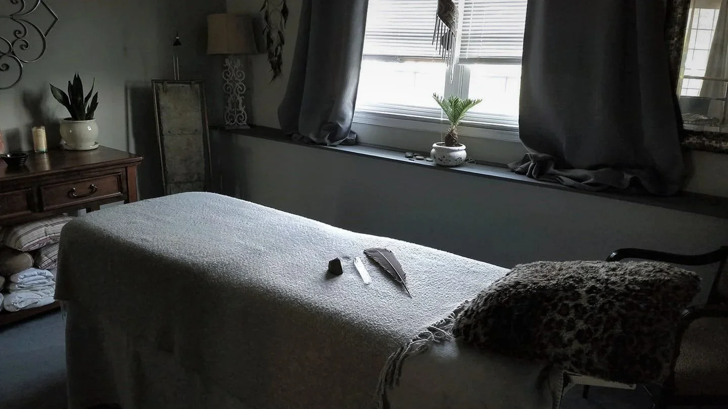 Cozy reiki studio with a comfortable reiki table, textured throw blanket, decorative pillows, houseplants, and curtained windows.