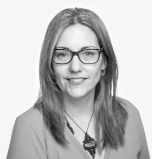 Black-and-white headshot of a smiling woman with long hair, glasses, and a pendant necklace