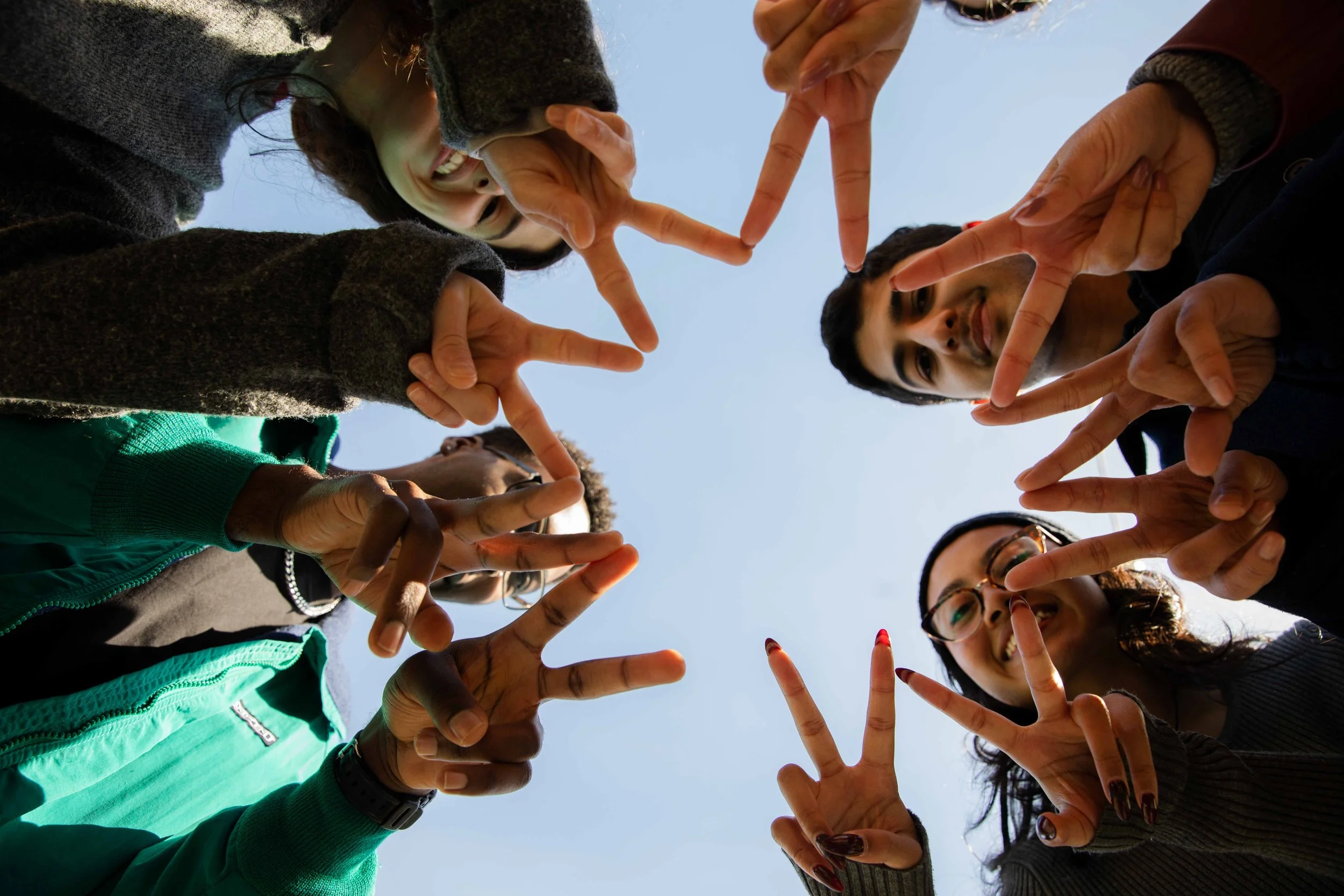 A group of young people standing close together outdoors, making peace signs with their hands and smiling, with a clear blue sky in the background.