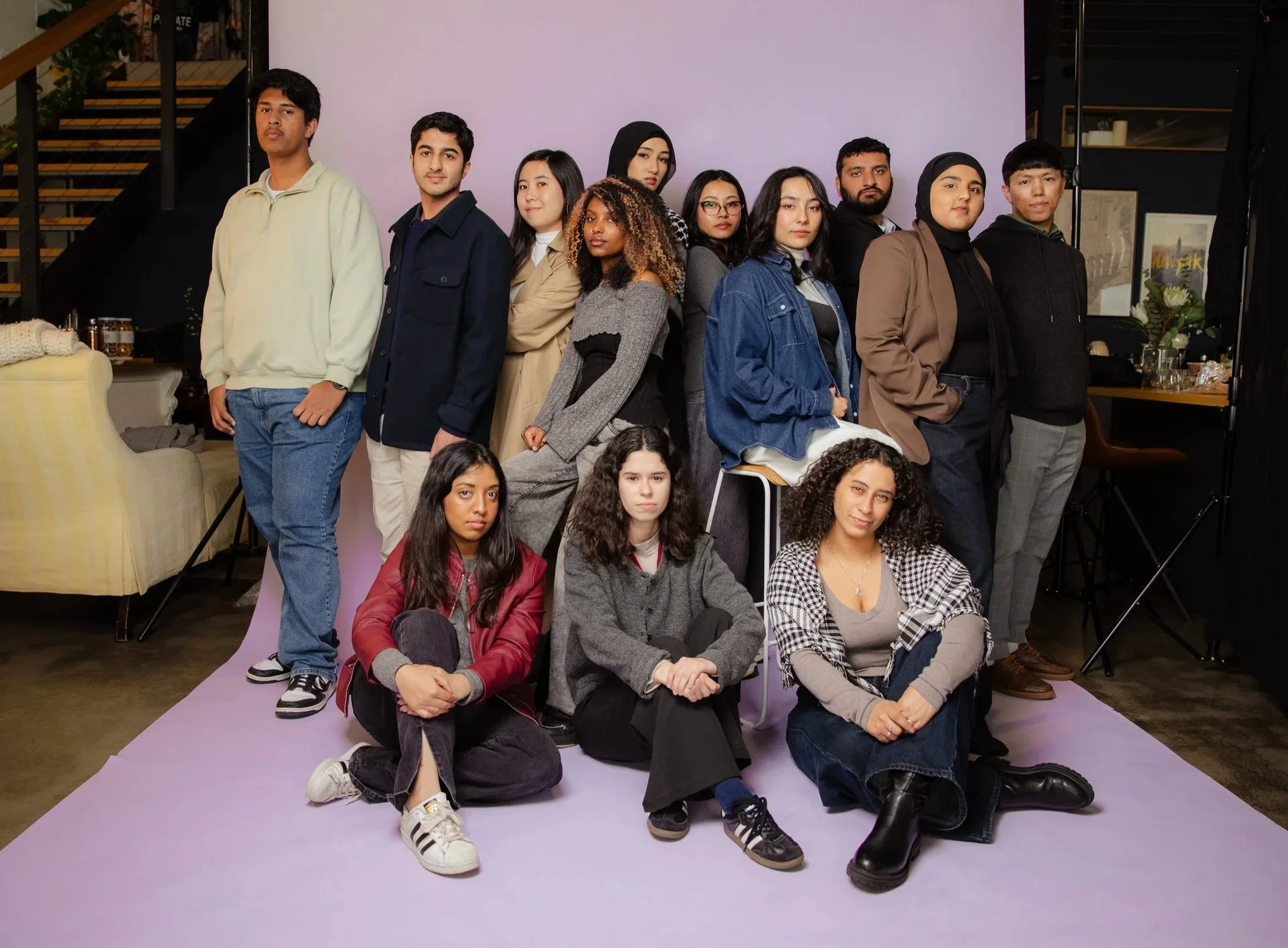 A diverse group of 13 young adults posing together in front of a purple backdrop, with some kneeling and others standing, inside a modern building with stairs and framed artwork in the background.