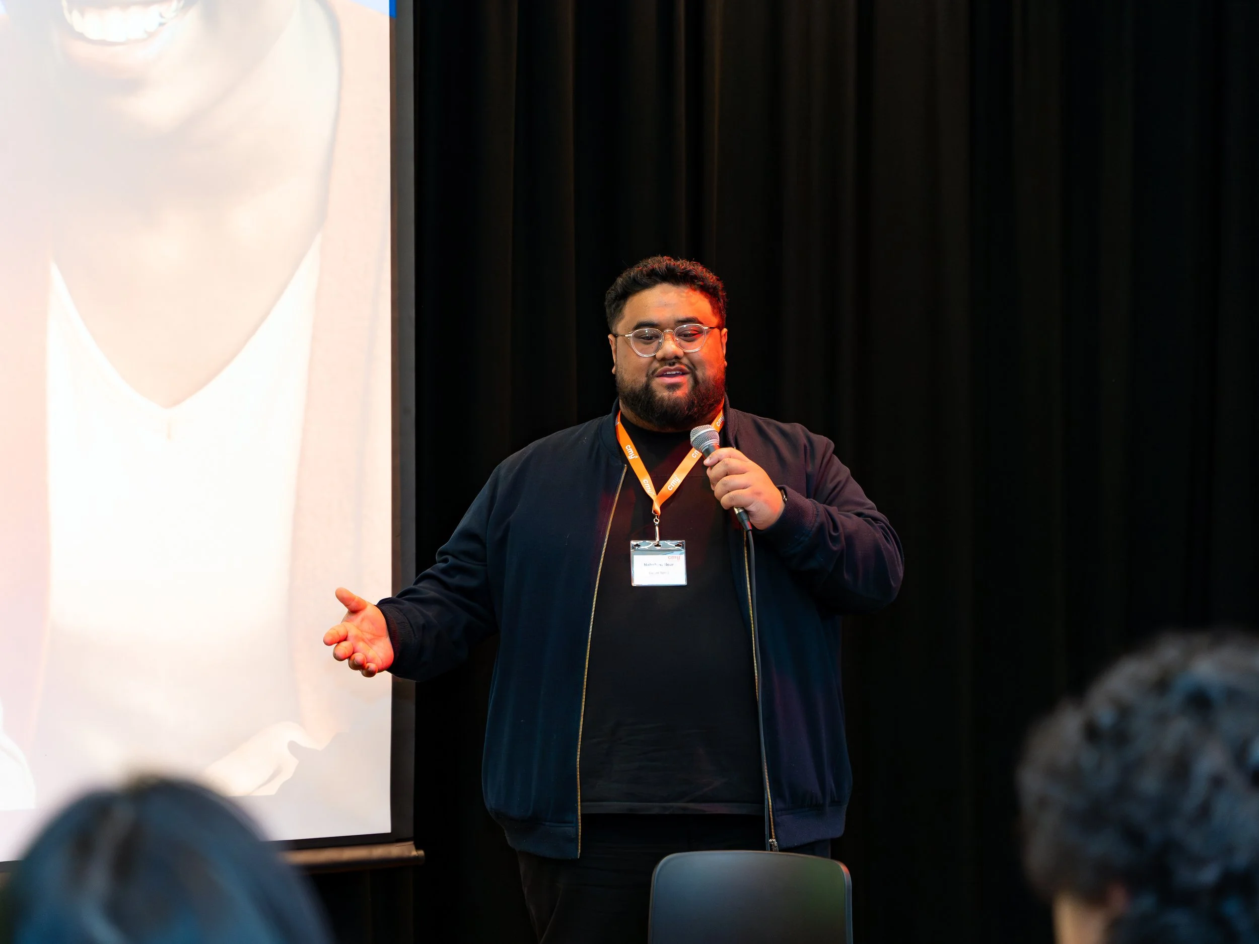 A man with glasses, a beard, and curly hair, wearing a black jacket and an orange lanyard, is speaking into a microphone while standing in front of a black curtain, with a large projection screen to his left.