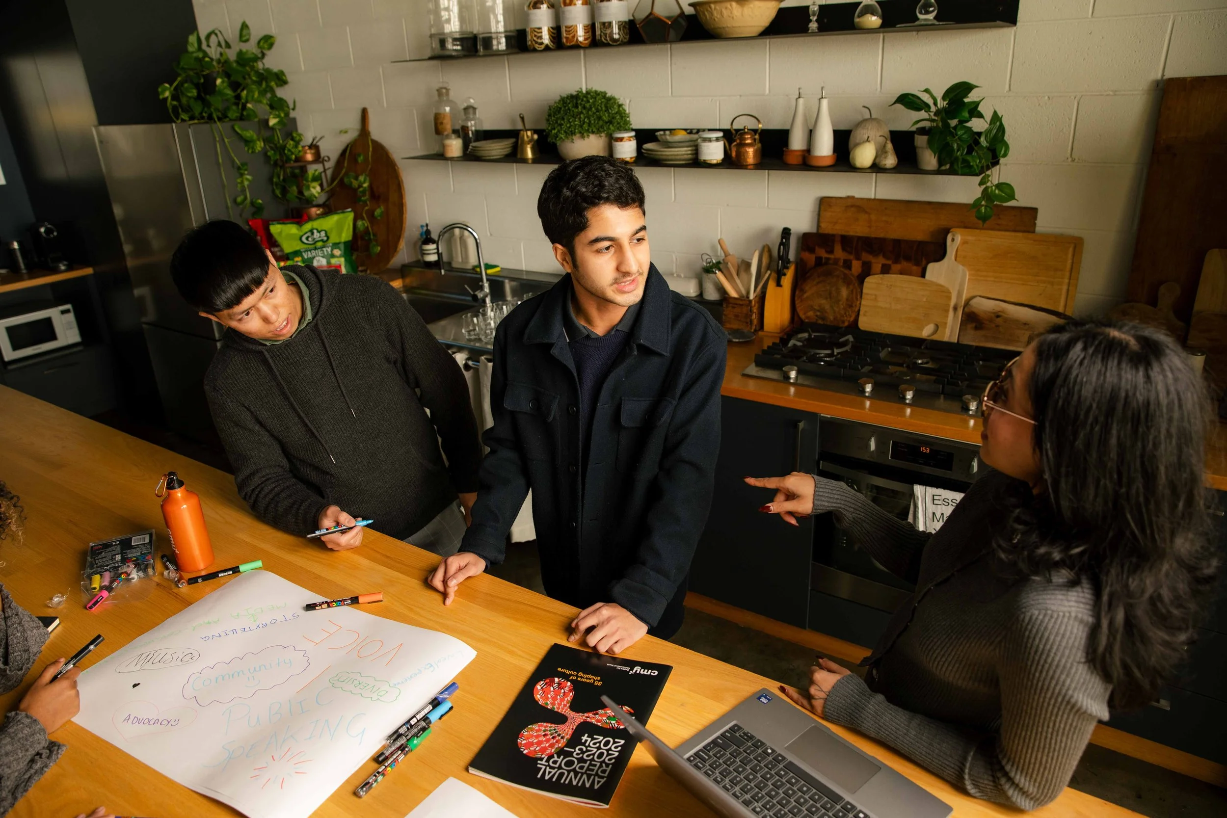 People gathered around a wooden table in a kitchen, engaging in a discussion. The table has markers, a water bottle, a laptop, and a poster with colorful writing related to community and advocacy. The kitchen has potted plants, shelves with dishes, and a stove in the background.
