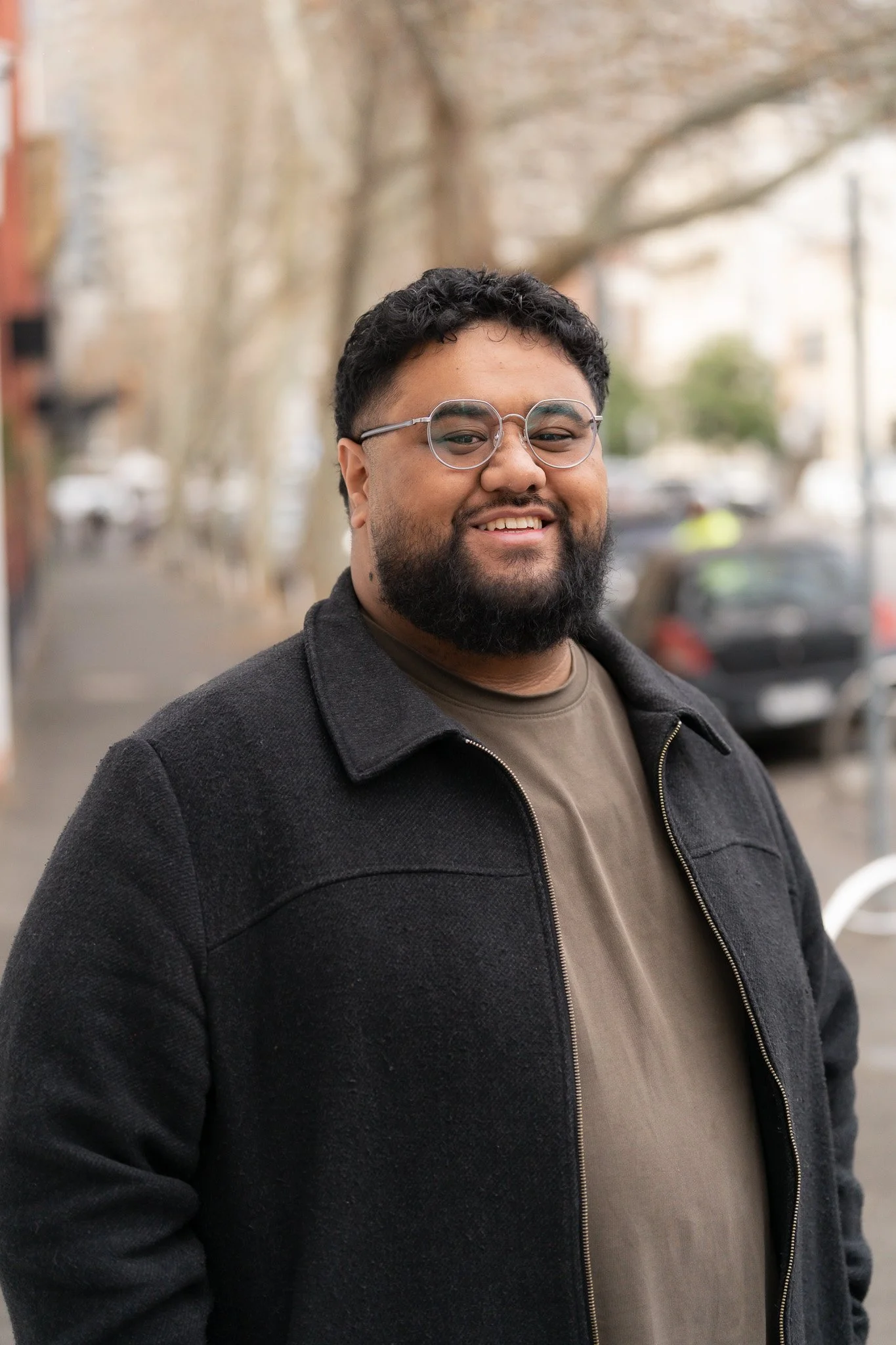 A smiling man with glasses and a beard stands outdoors on a city street, with trees and parked cars in the background.