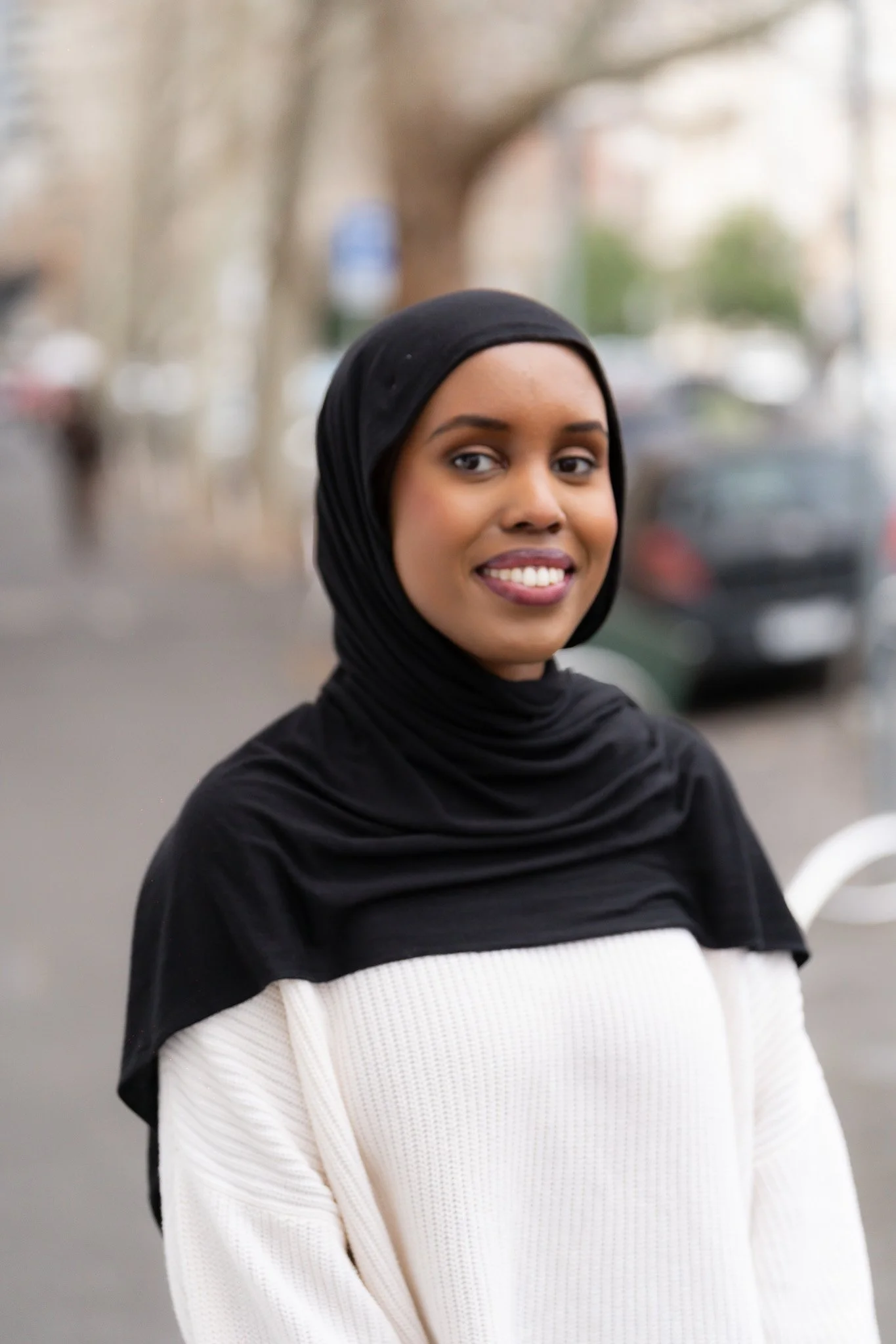 A young woman with a headscarf smiling outdoors on a city street.