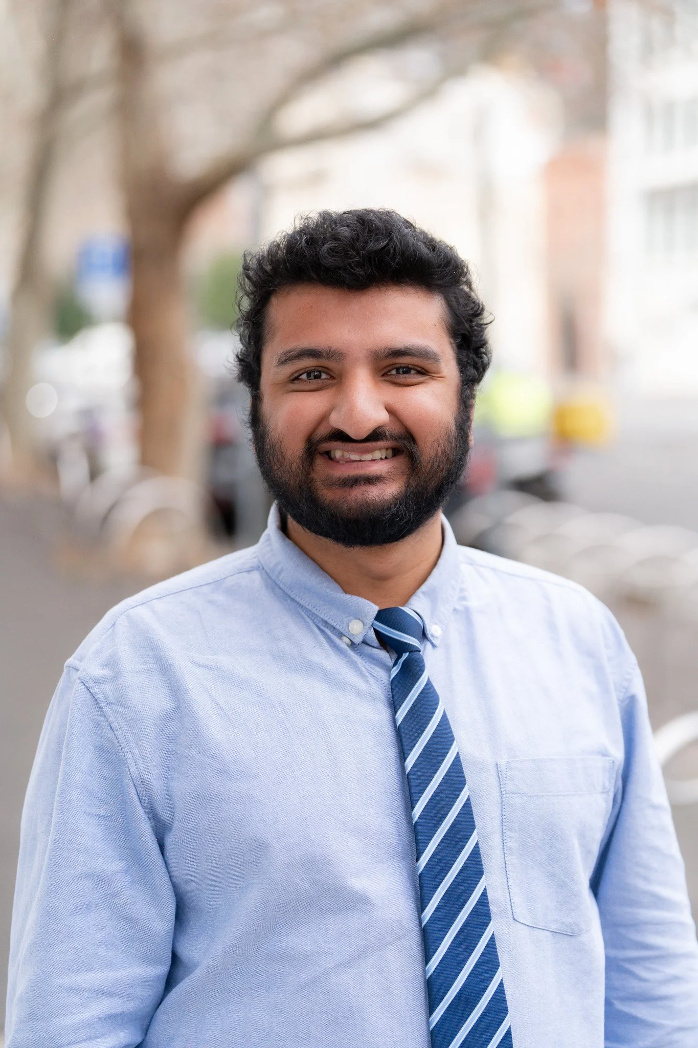 Smiling man in blue dress shirt and striped tie standing outdoors amid blurred city background with trees and cars.