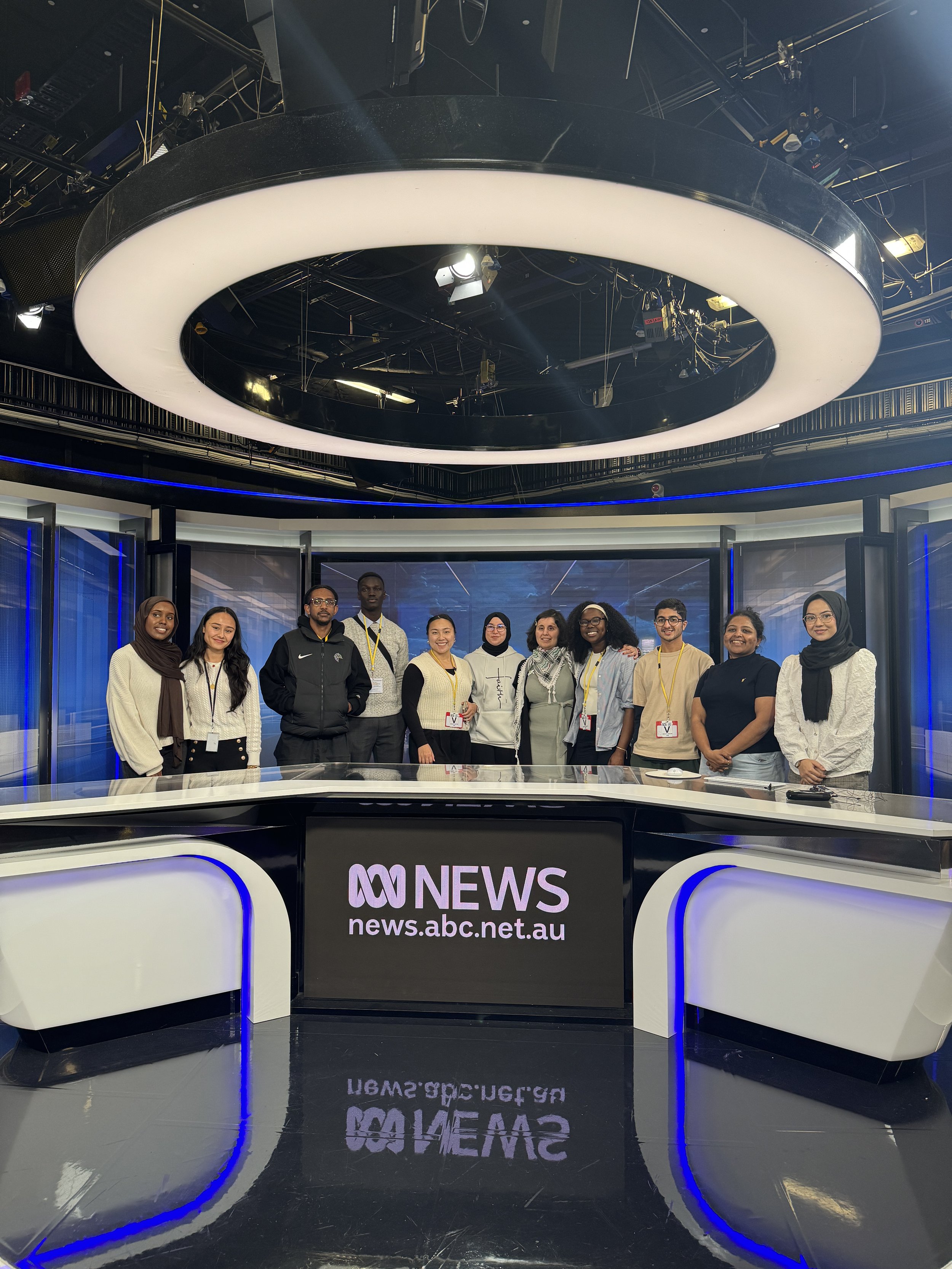 A group of diverse journalists and news staff posing in a television studio behind a news desk with an ABC News logo and website, under a large circular ceiling light.