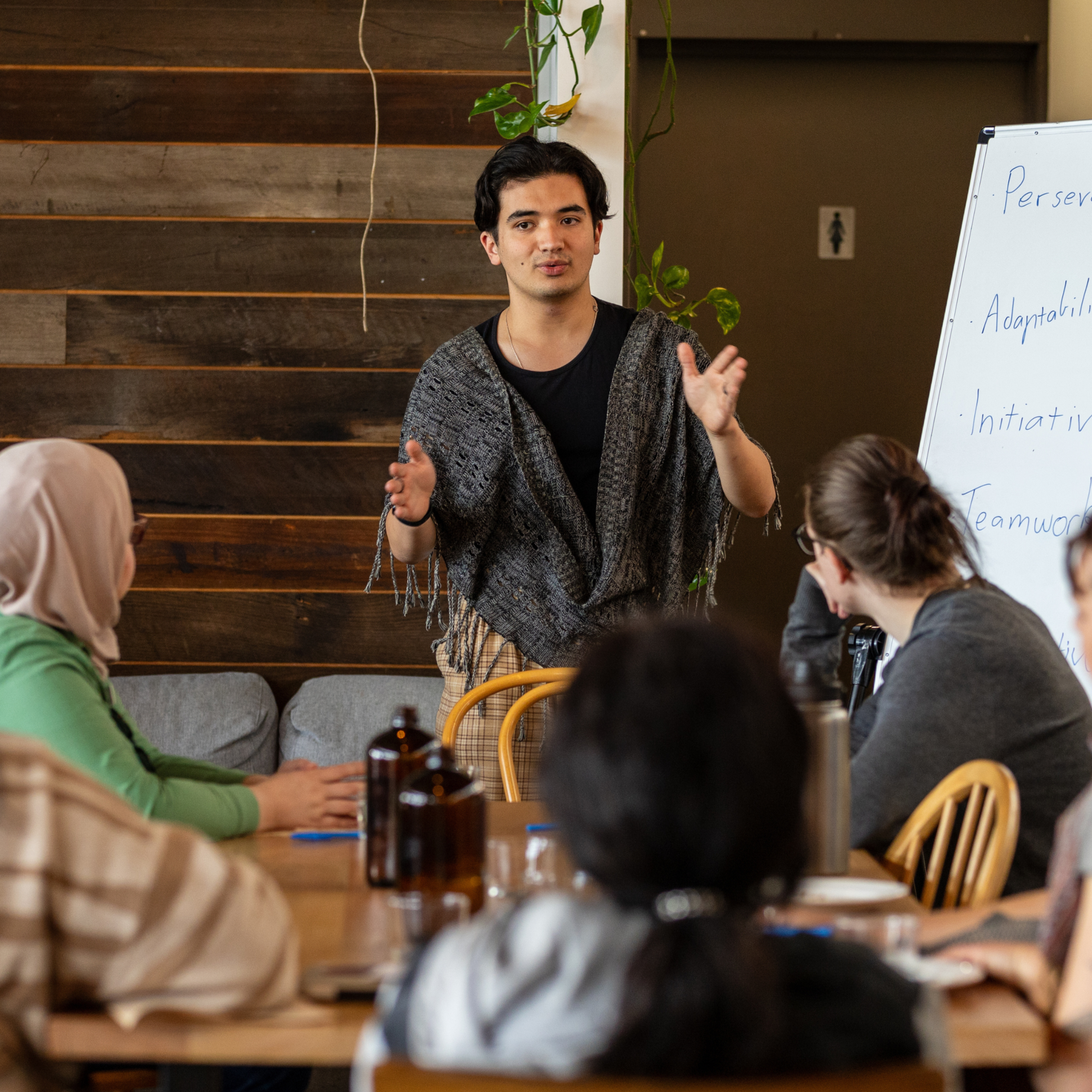 A man is speaking to a group of people in a room, standing near a whiteboard with notes. The audience includes a woman in a hijab and others. The room has wood-paneled walls and hanging green plants.