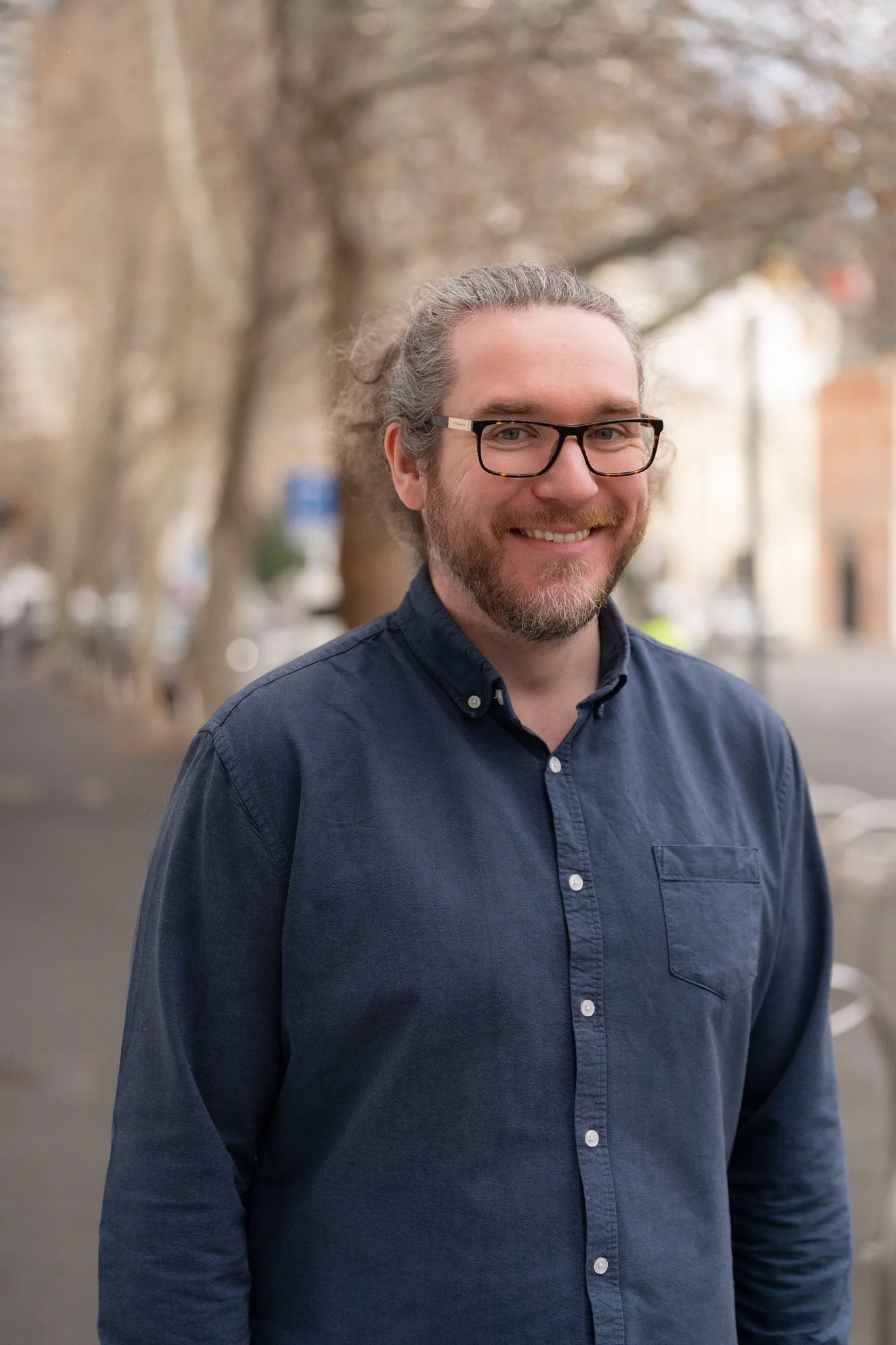 A man with gray hair, glasses, and a beard smiling outdoors in front of trees and buildings, wearing a dark blue button-up shirt.