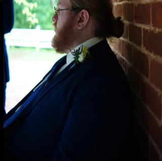 A man with red hair and a beard wearing glasses and a dark suit, sitting against a brick wall.