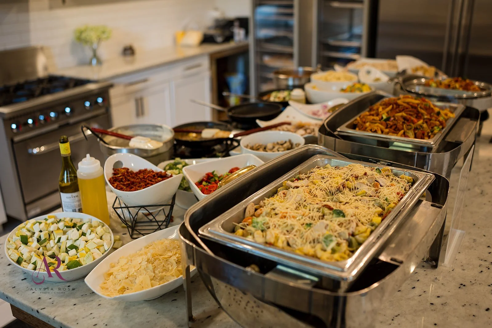 A buffet table with various dishes including pasta, vegetables, salads, and toppings, set in a kitchen with white cabinets and appliances.