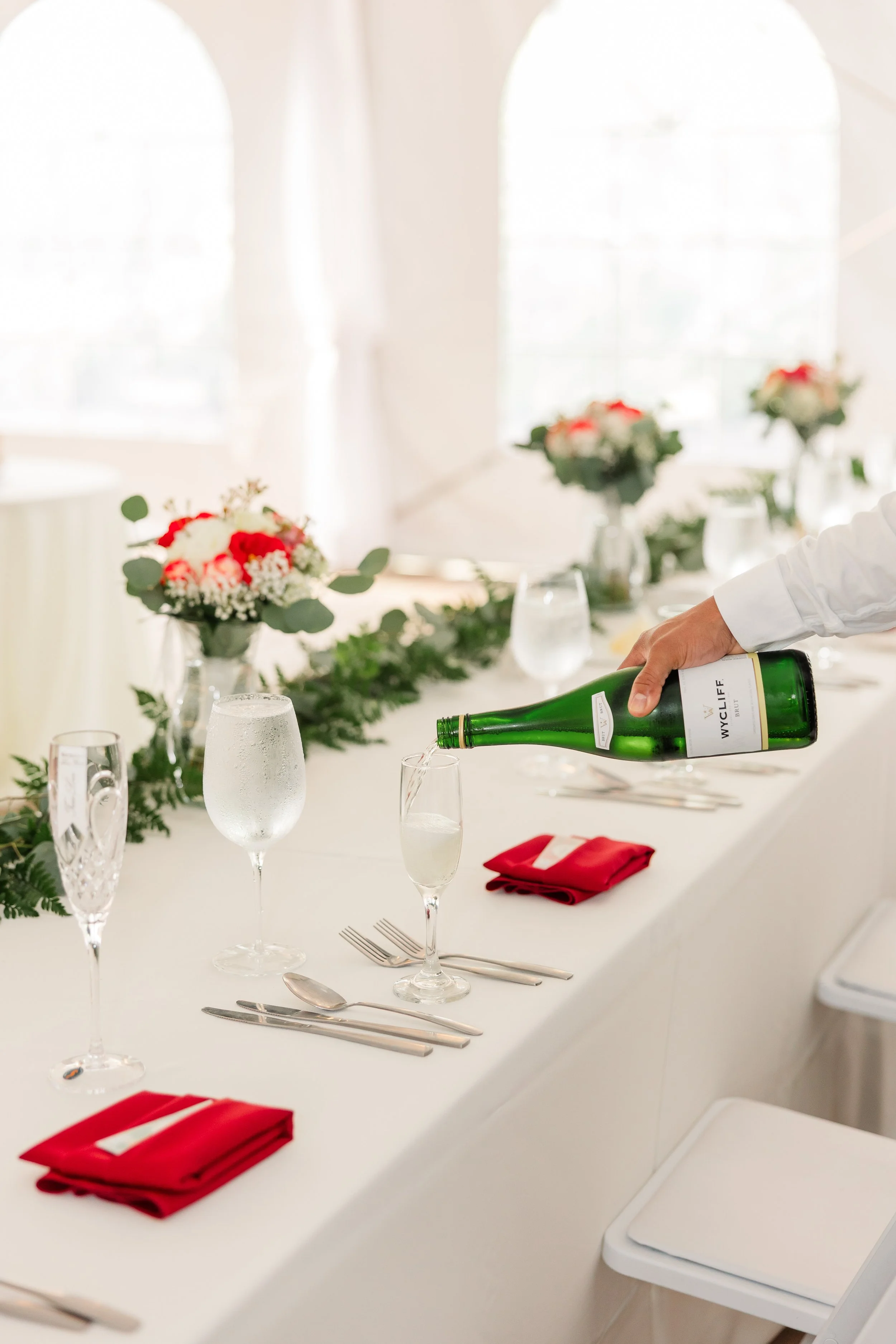 Elegant banquet table set with silverware, glasses, red napkins, and floral centerpieces, with a person pouring wine.