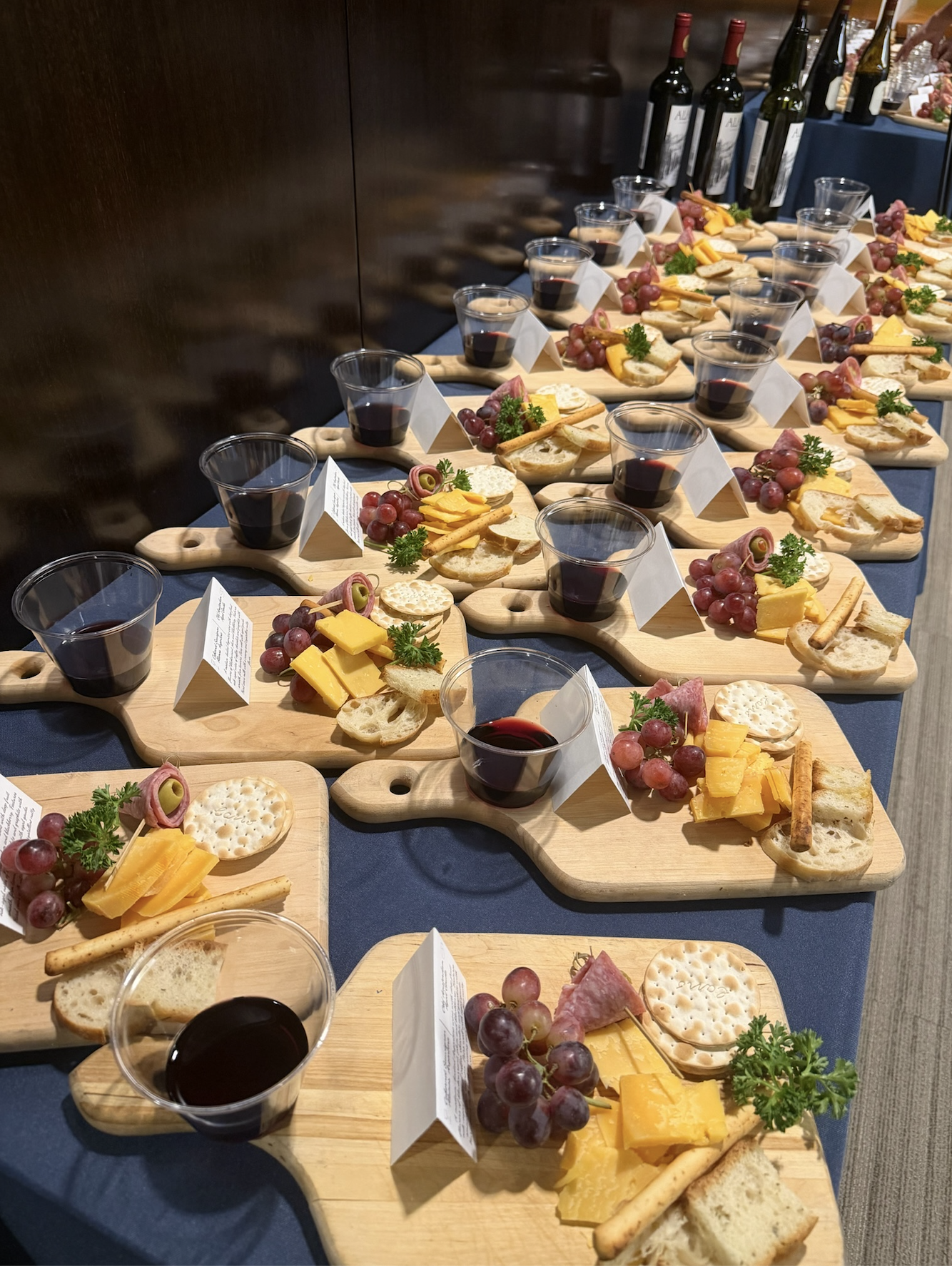Multiple wooden serving boards with cheese, grapes, crackers, and a glass of red wine arranged on a dark table at a buffet table.