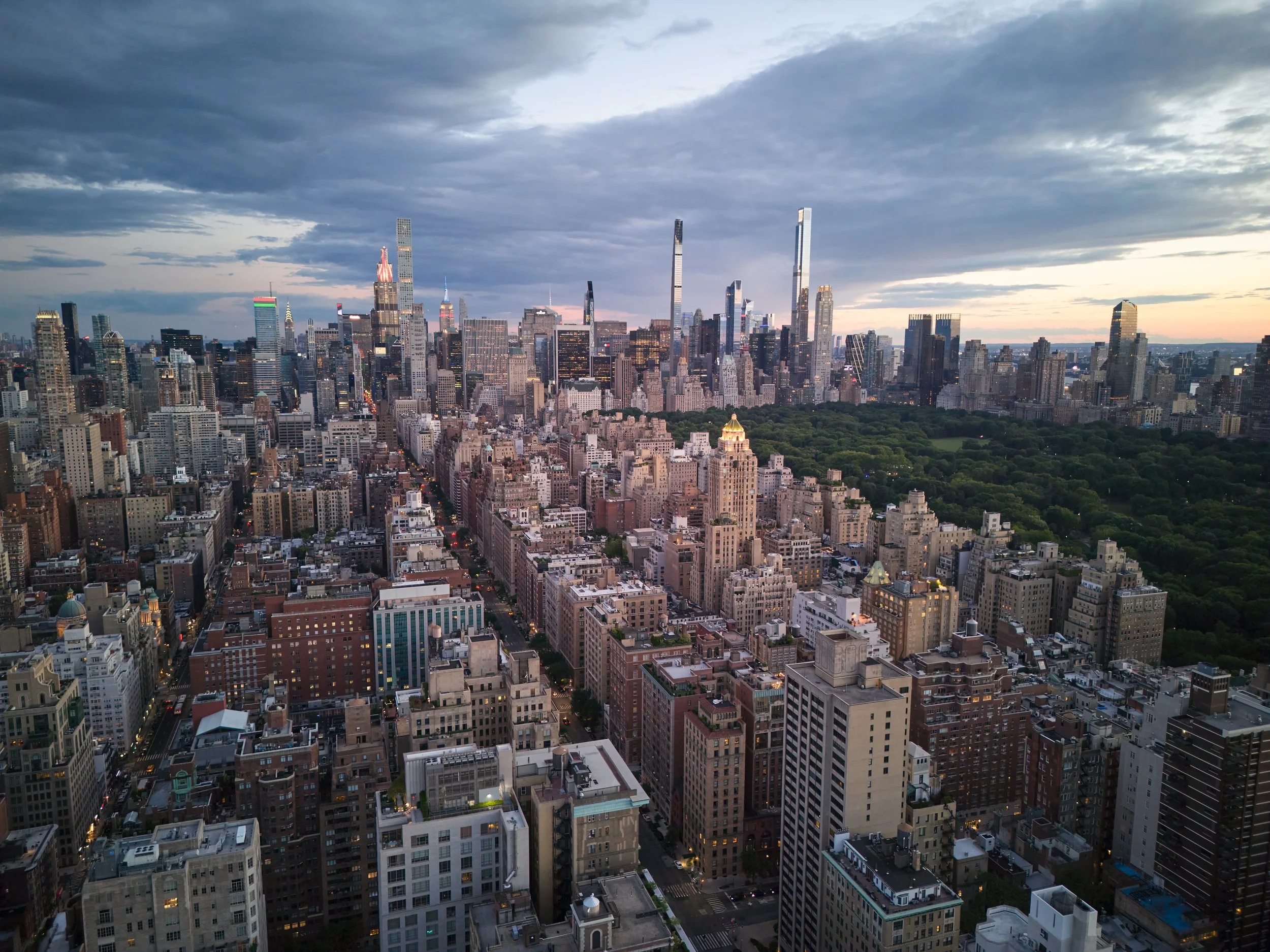 A city skyline featuring numerous tall skyscrapers under a partly cloudy sky, with some buildings illuminated, and a green park in the foreground.