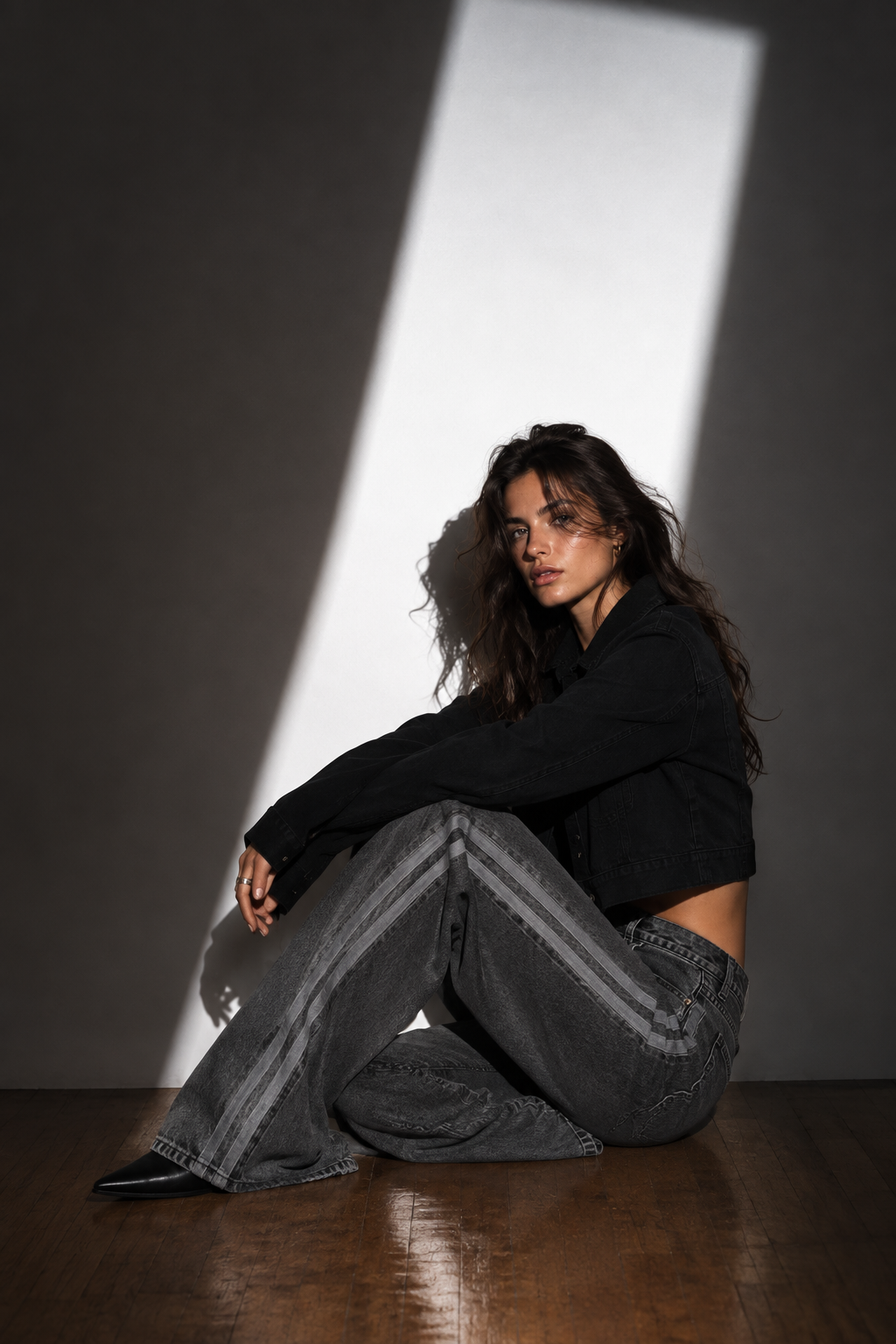 A young woman with long wavy brown hair, wearing a black denim jacket and loose gray stripe pants, sits on the floor against a wall with a bright light creating a shadow behind her.