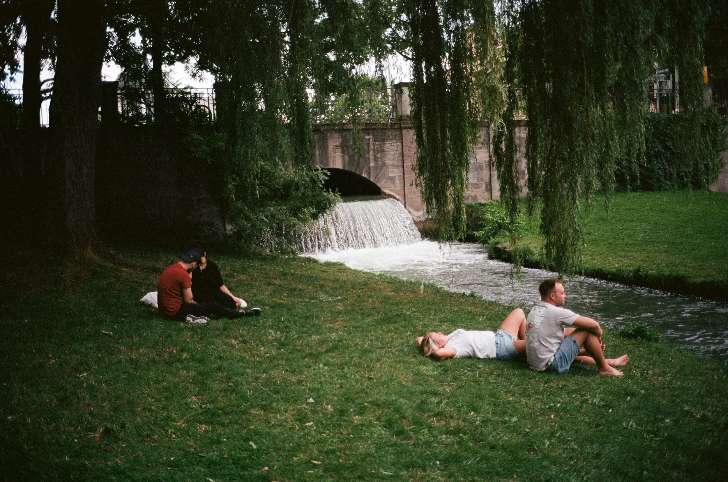People relaxing on grass near a small waterfall and a stone bridge, surrounded by trees.