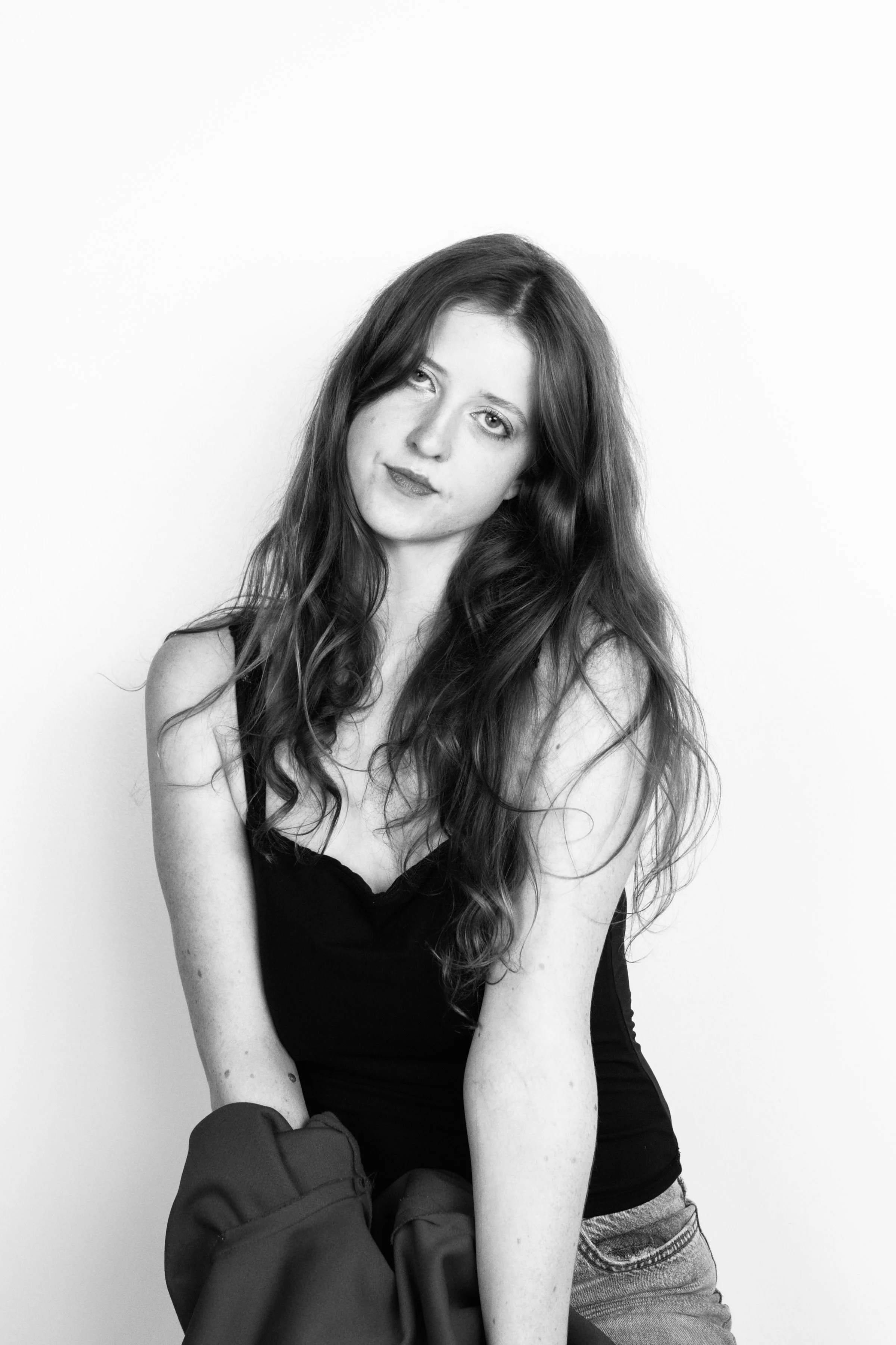 Black and white portrait of a young woman with long, wavy hair, wearing a sleeveless top and sitting with crossed arms, against a plain background.