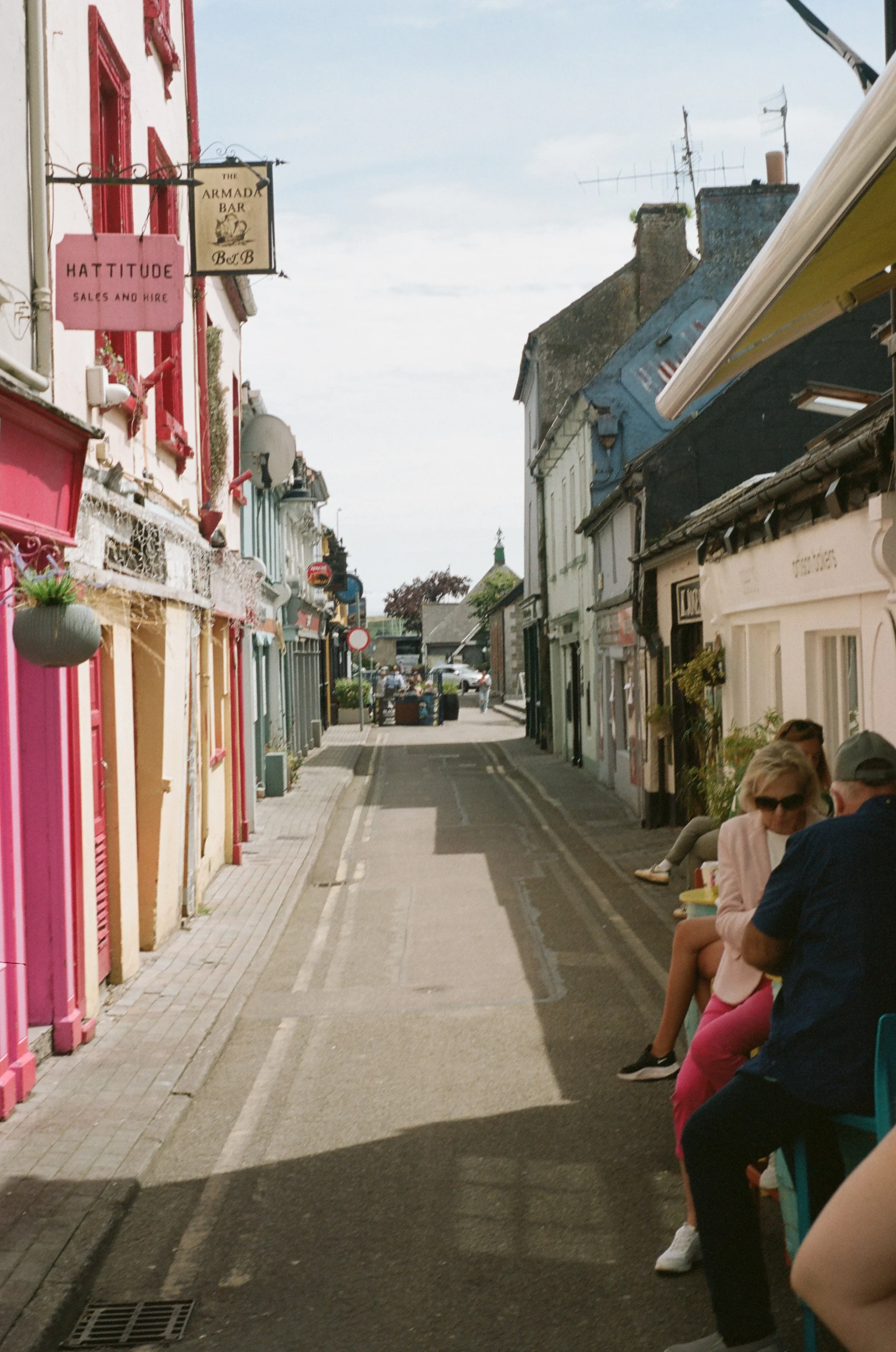 A street scene with colorful buildings on either side, people sitting on a bench outside, and a cloudy sky overhead.