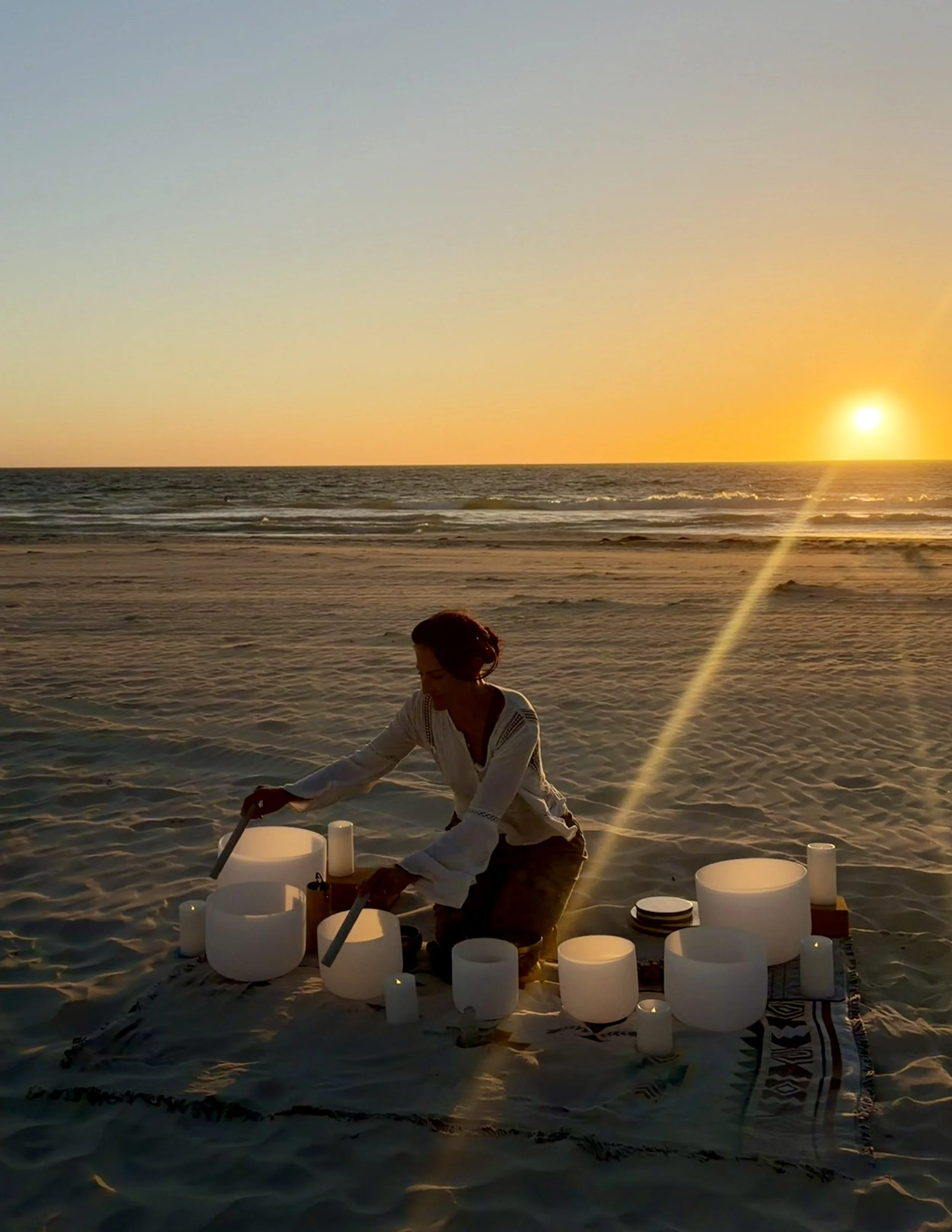 A woman is sitting on a beach at sunset, surrounded by white candles, with the ocean and setting sun in the background.