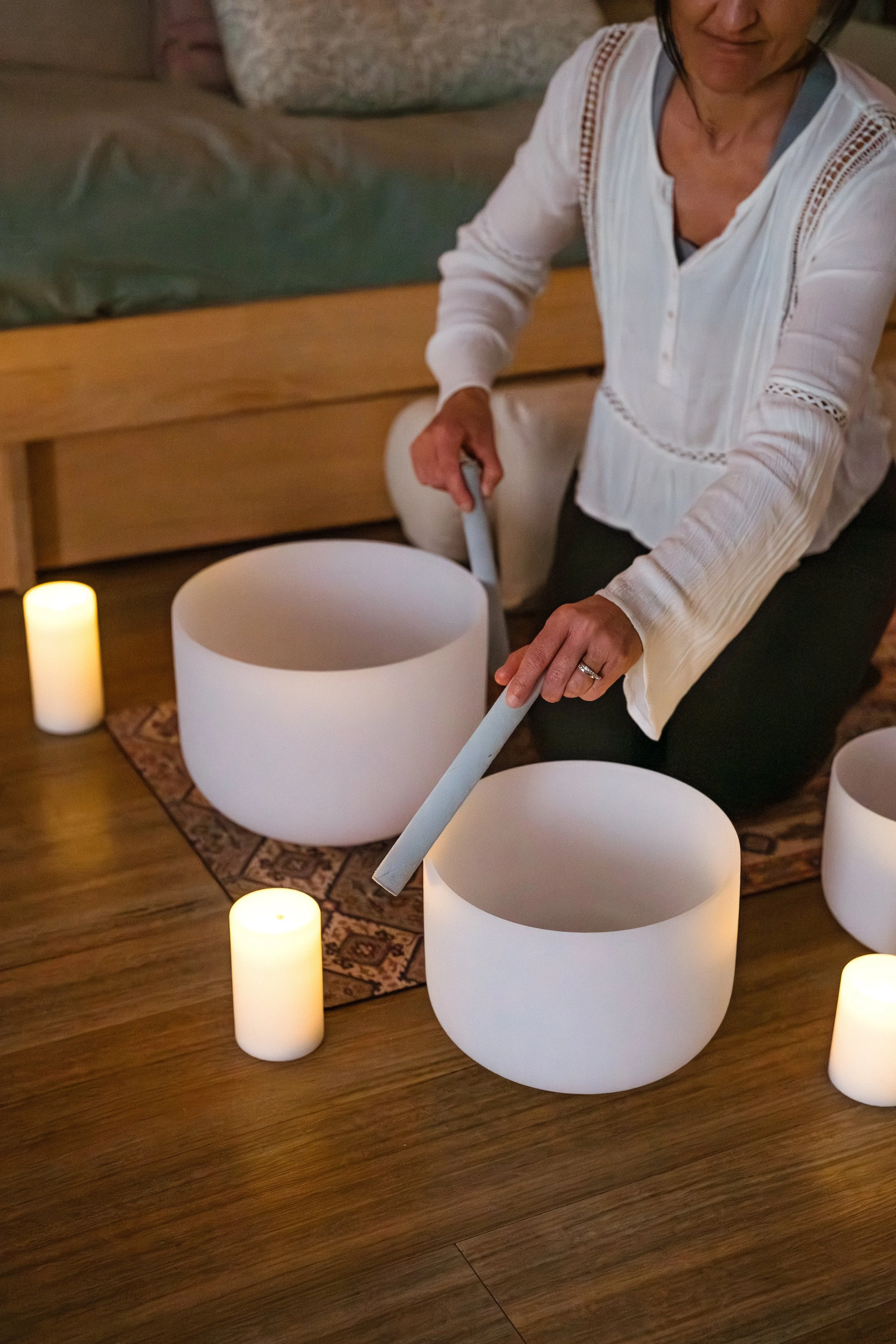 A woman playing a white crystal sound bowl surrounded by candles on a wooden floor.