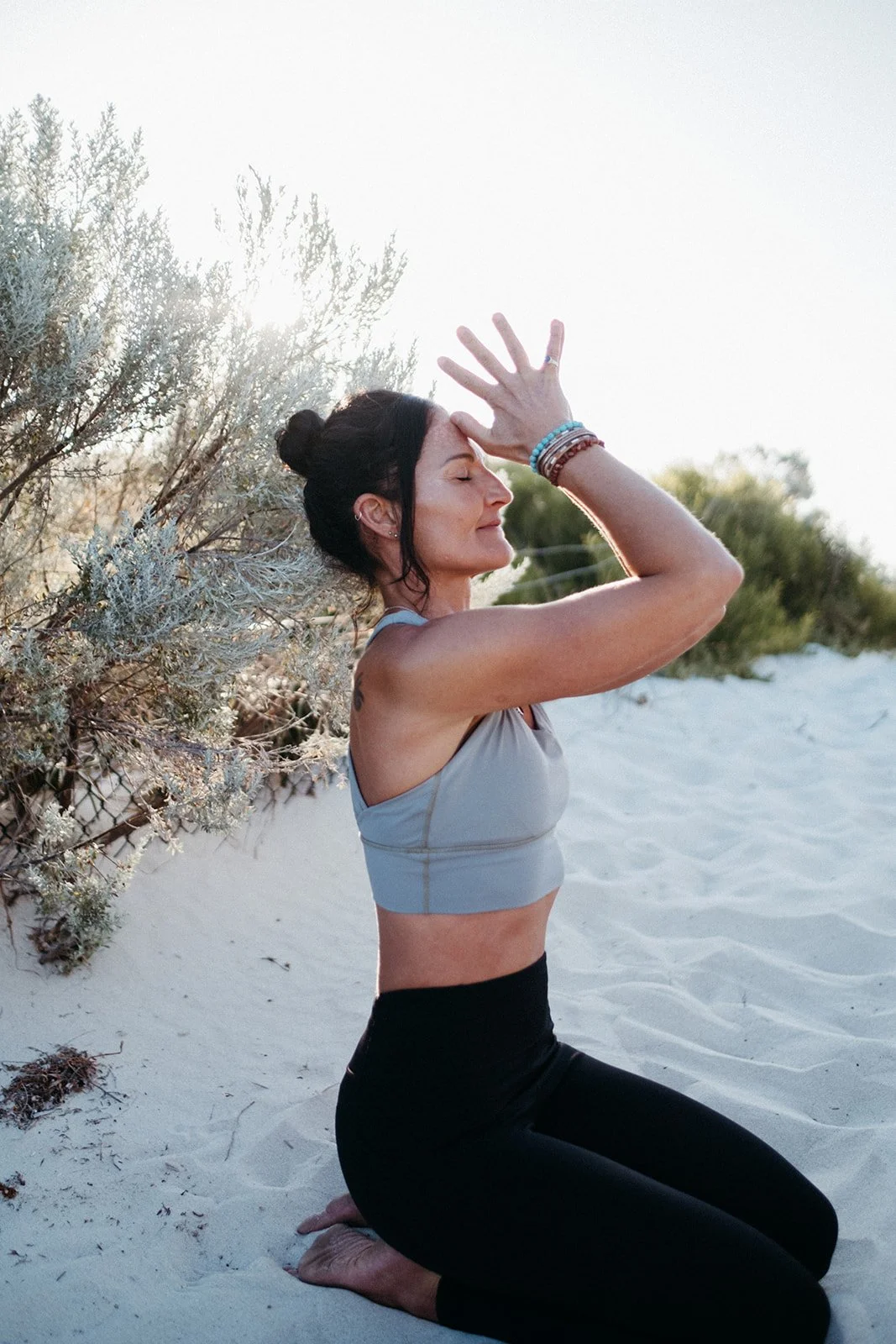 A woman practicing yoga outdoors on sandy ground; she is kneeling with her eyes closed, hands raised above her forehead, surrounded by bushes and clear sky.
