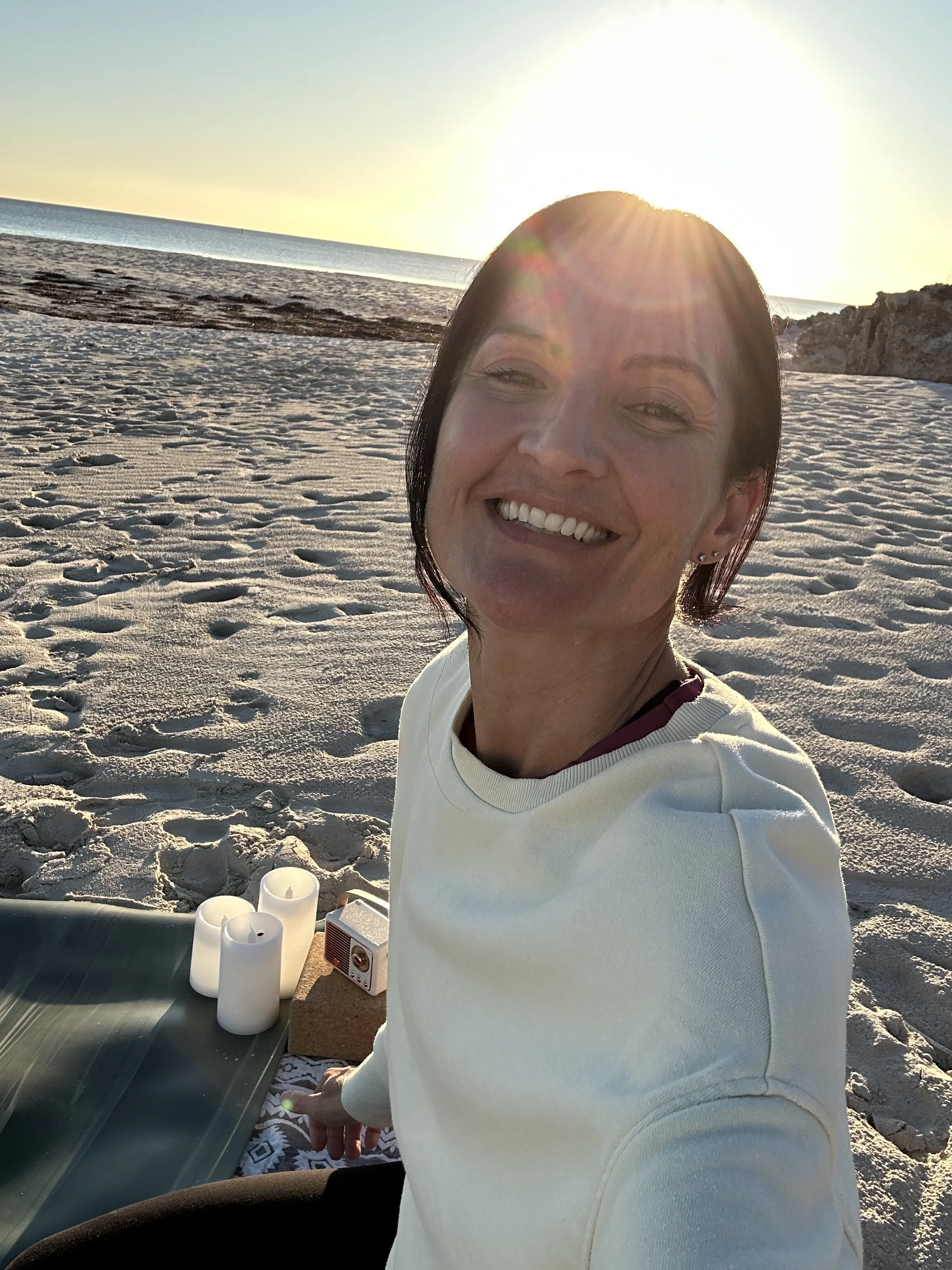 A woman taking a selfie at the beach during sunset, with sand, rocks, and the ocean in the background. On the table in front of her are white candles and a small speaker.