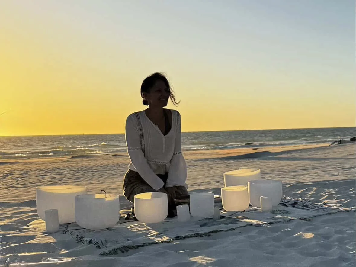 A woman sits on a blanket on a sandy beach during sunset, surrounded by white singing bowls.