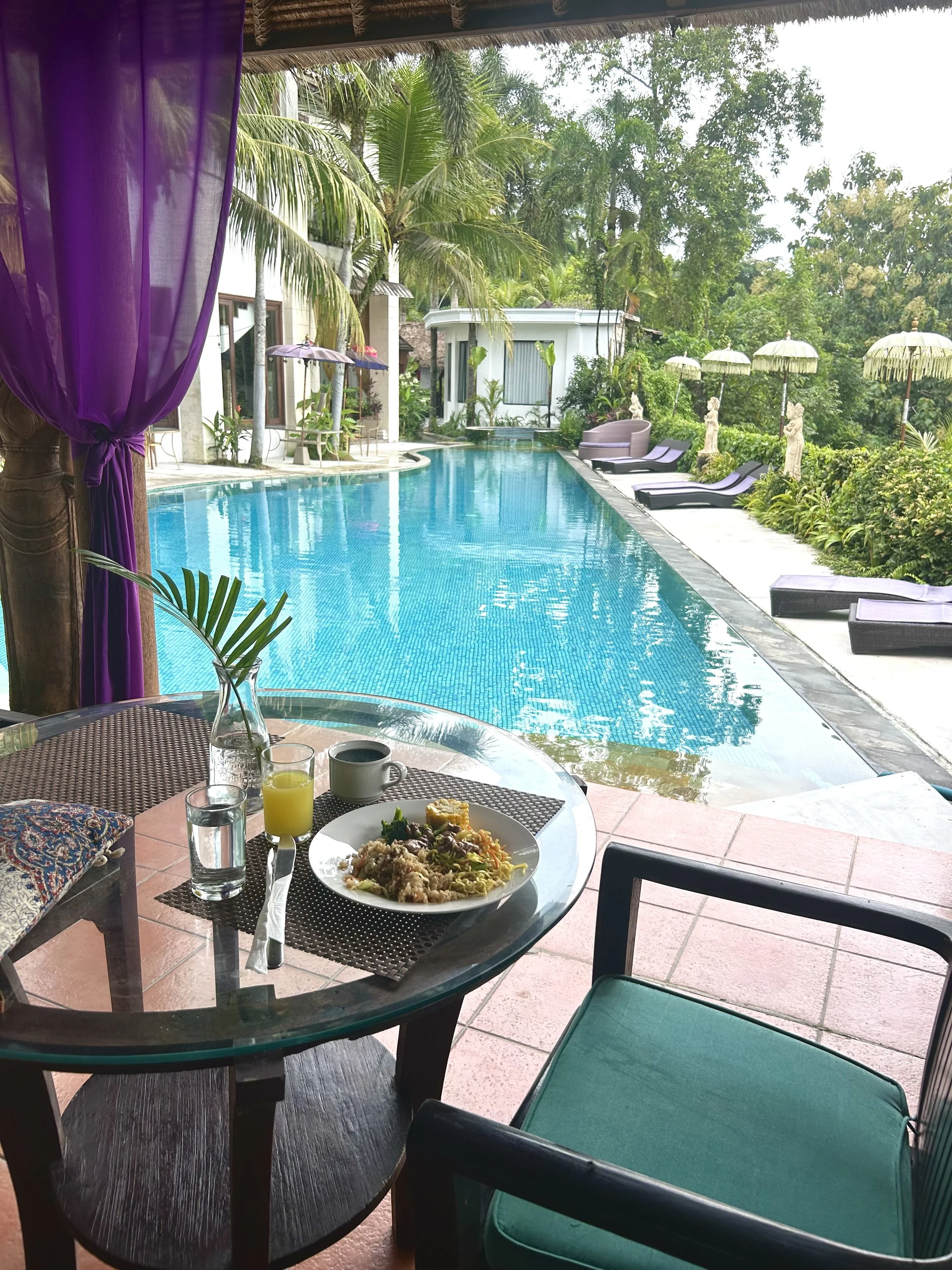A table with breakfast food, including a plate of rice, vegetables, and eggs, along with a glass of orange juice and a mug on a balcony overlooking a swimming pool surrounded by lounge chairs, umbrellas, and tropical plants.