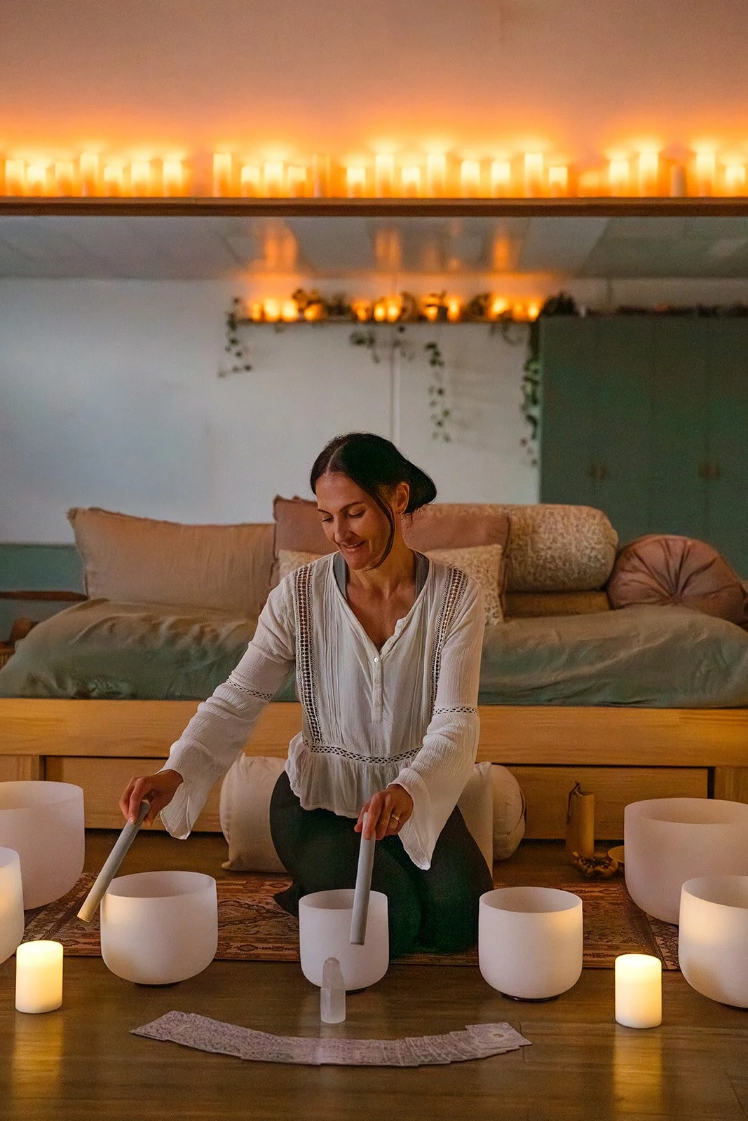 A woman kneeling on the floor surrounded by lit candles, playing crystal singing bowls in a cozy, softly lit room.