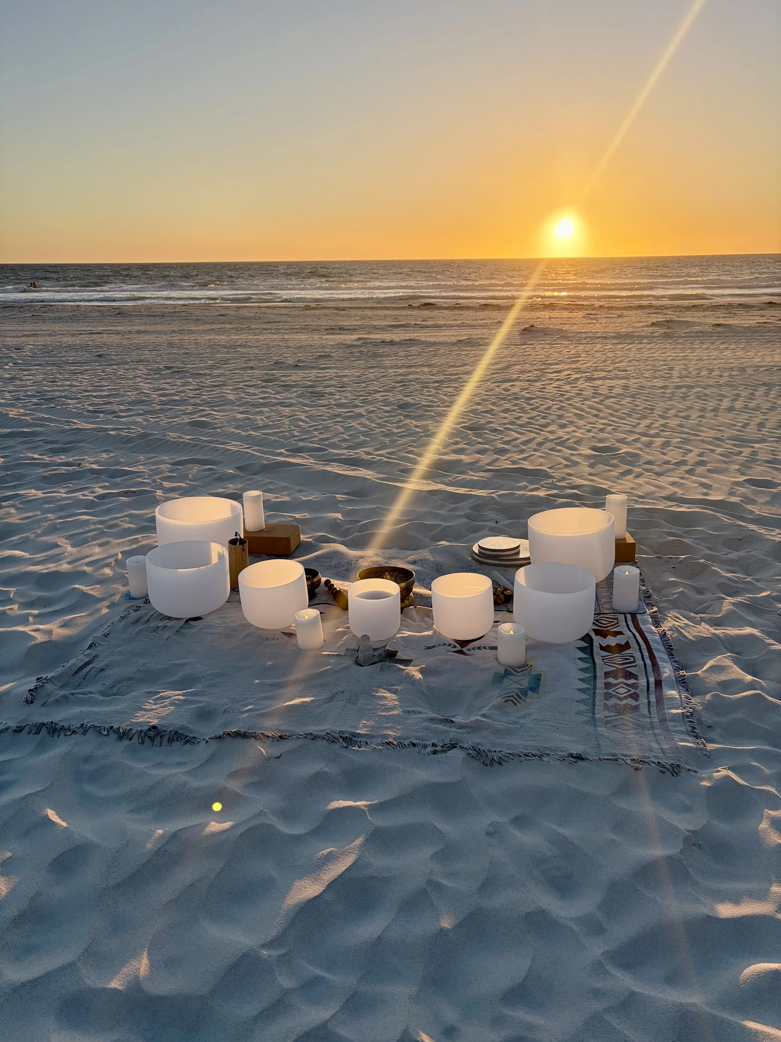 A beach scene during sunset with white singing bowls and candles arranged on a rug in the sand, ocean waves in the background, and the sun low in the sky.