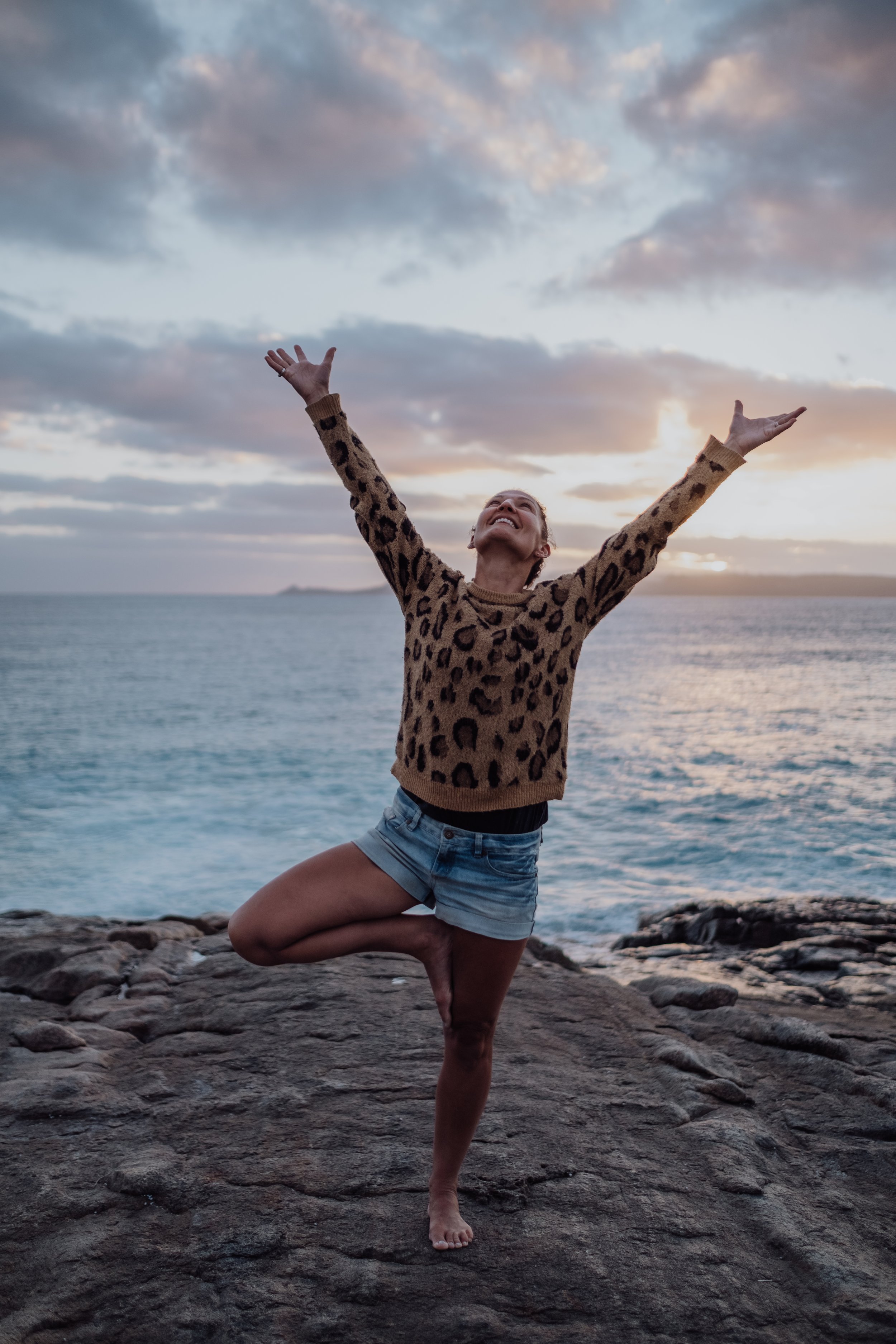 A woman practicing yoga outdoors near a body of water during sunset, balancing on one leg with arms raised, wearing a leopard print sweater and denim shorts.