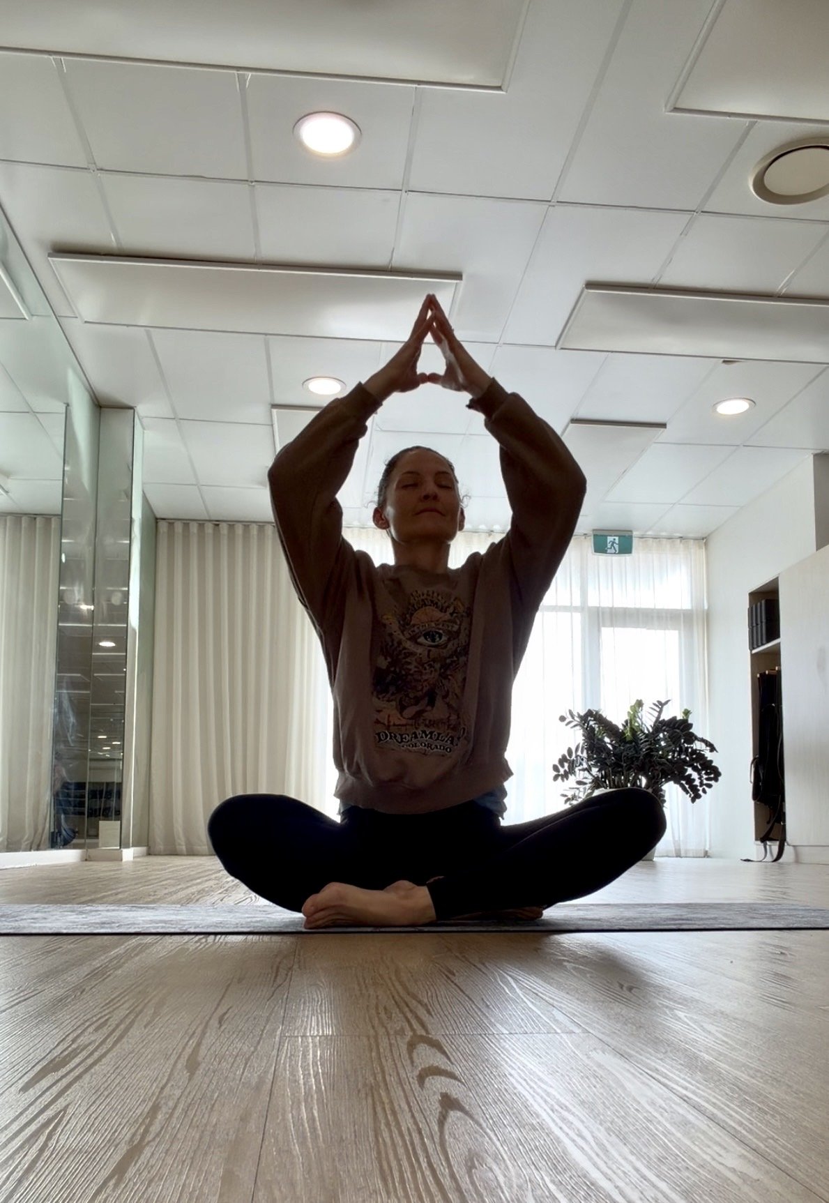 A woman practicing yoga indoors, sitting cross-legged on a mat, with her hands in a prayer position raised above her head, in a bright room with large windows, curtains, and a potted plant.
